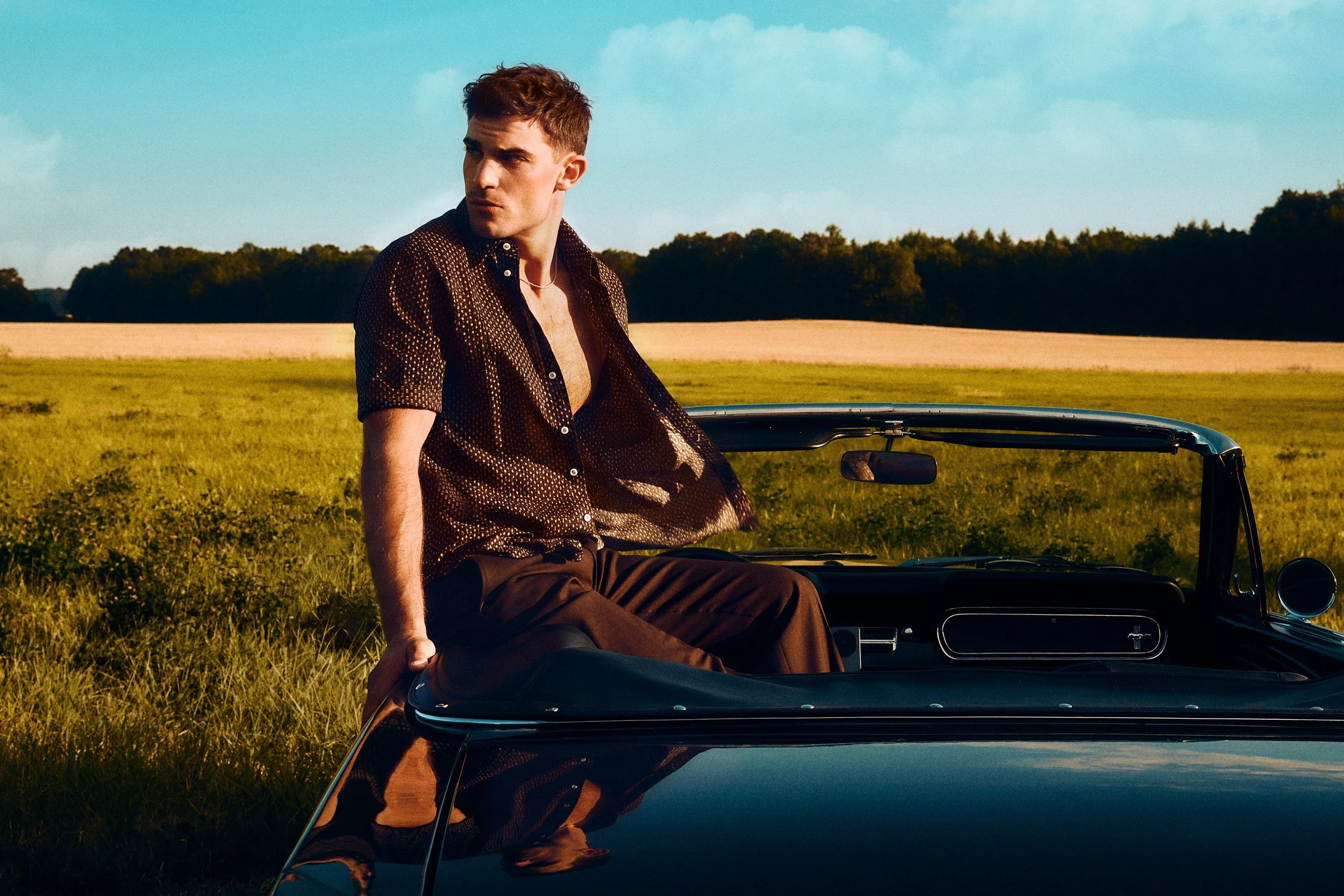 Man sitting on the side of a convertible in a dark open shirt and brown trousers.