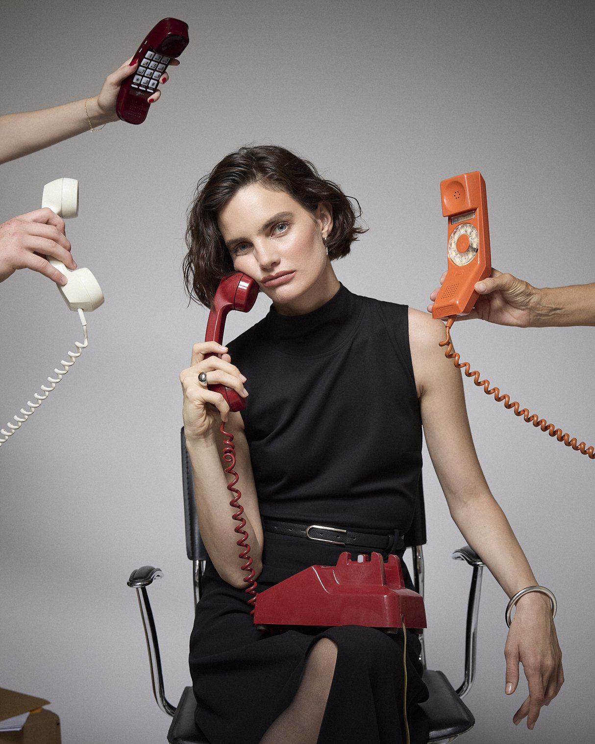 Woman in a black dress holding a red telephone as several other receivers are held around her.