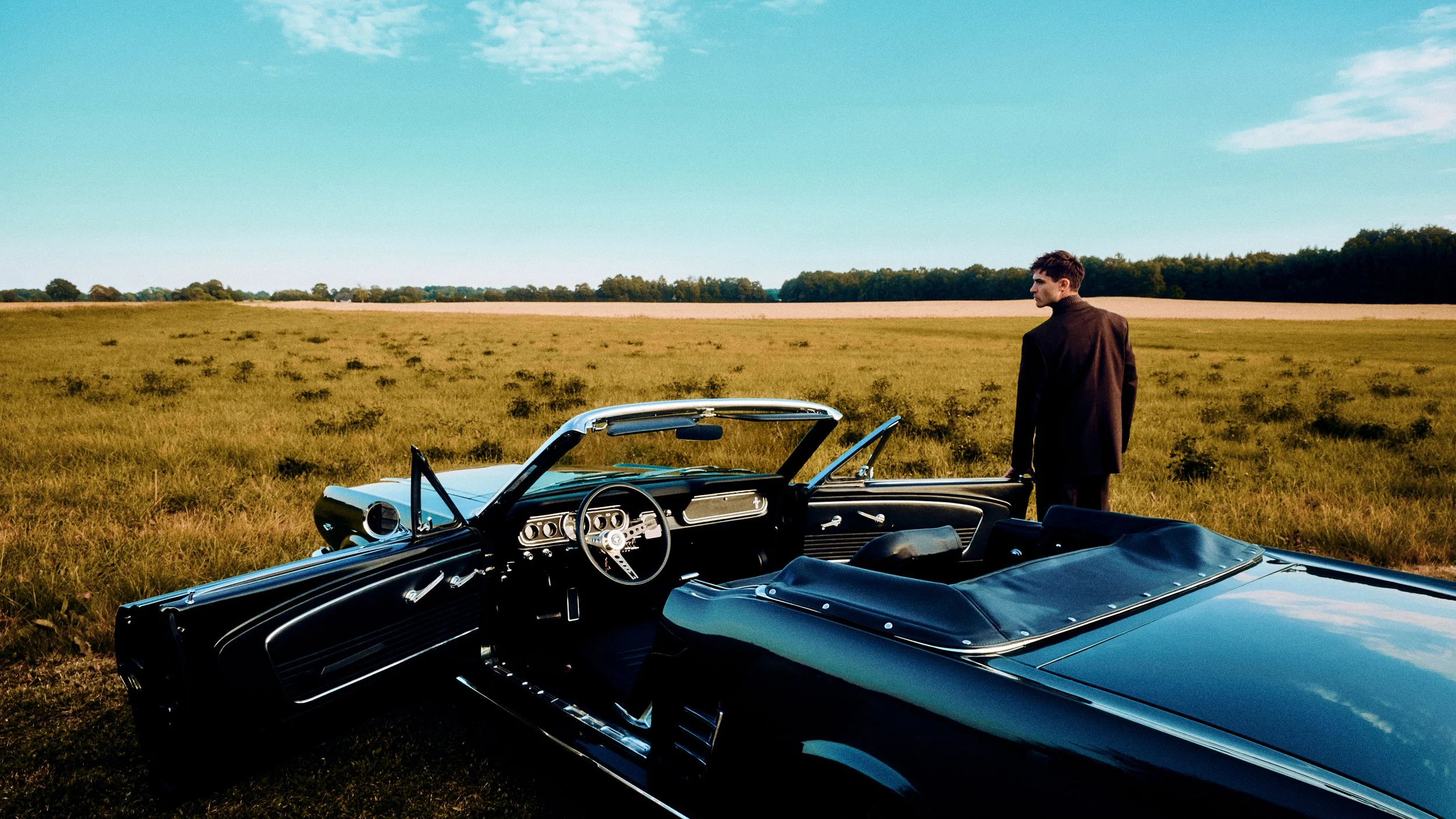 Wide view of a turquoise convertible and a man standing beside it in the countryside.