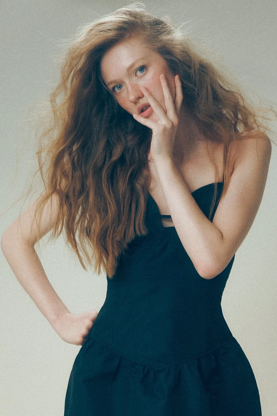 Woman in a black dress leaning forward with one hand against her cheek.