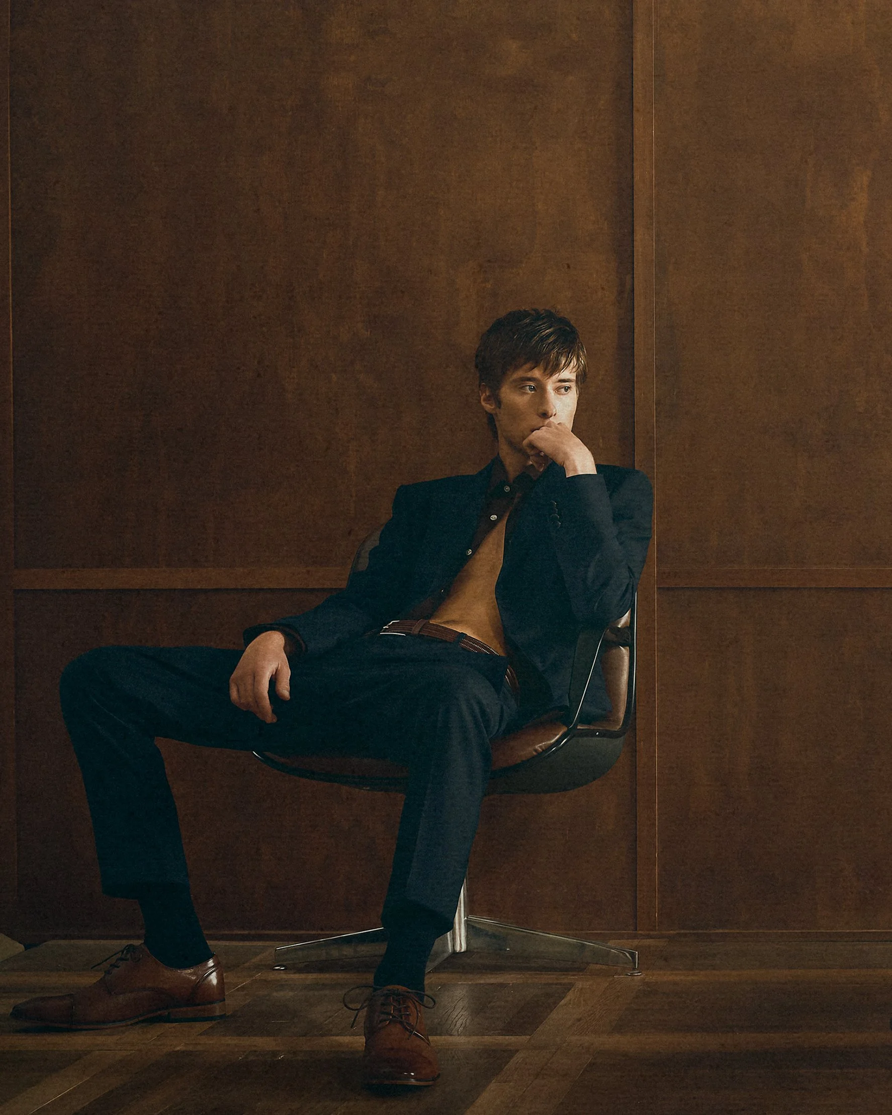 Man in a dark suit seated against wood panelling with one hand beside his face.