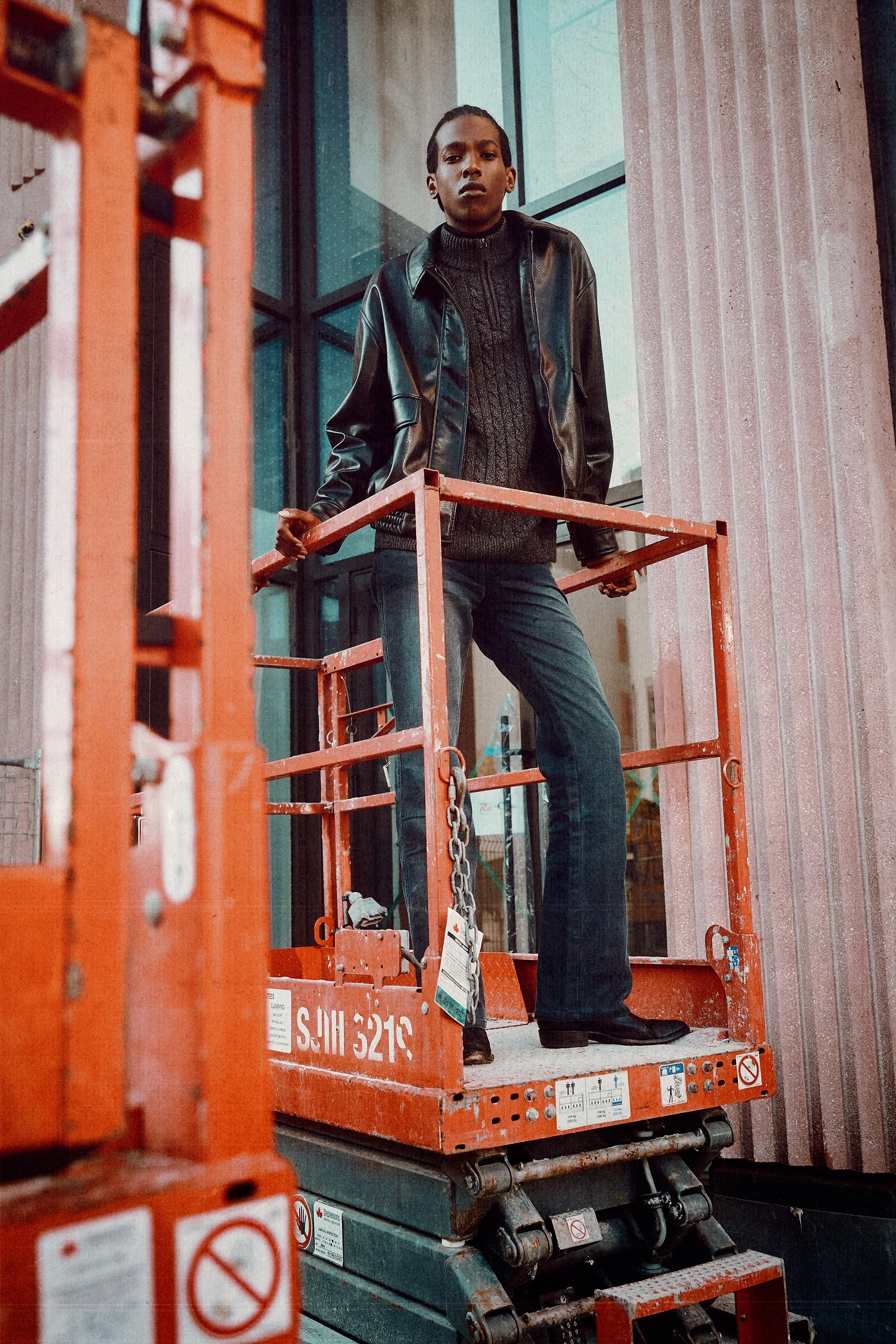 Man in a dark leather jacket, knit sweater and jeans standing on an orange construction lift.