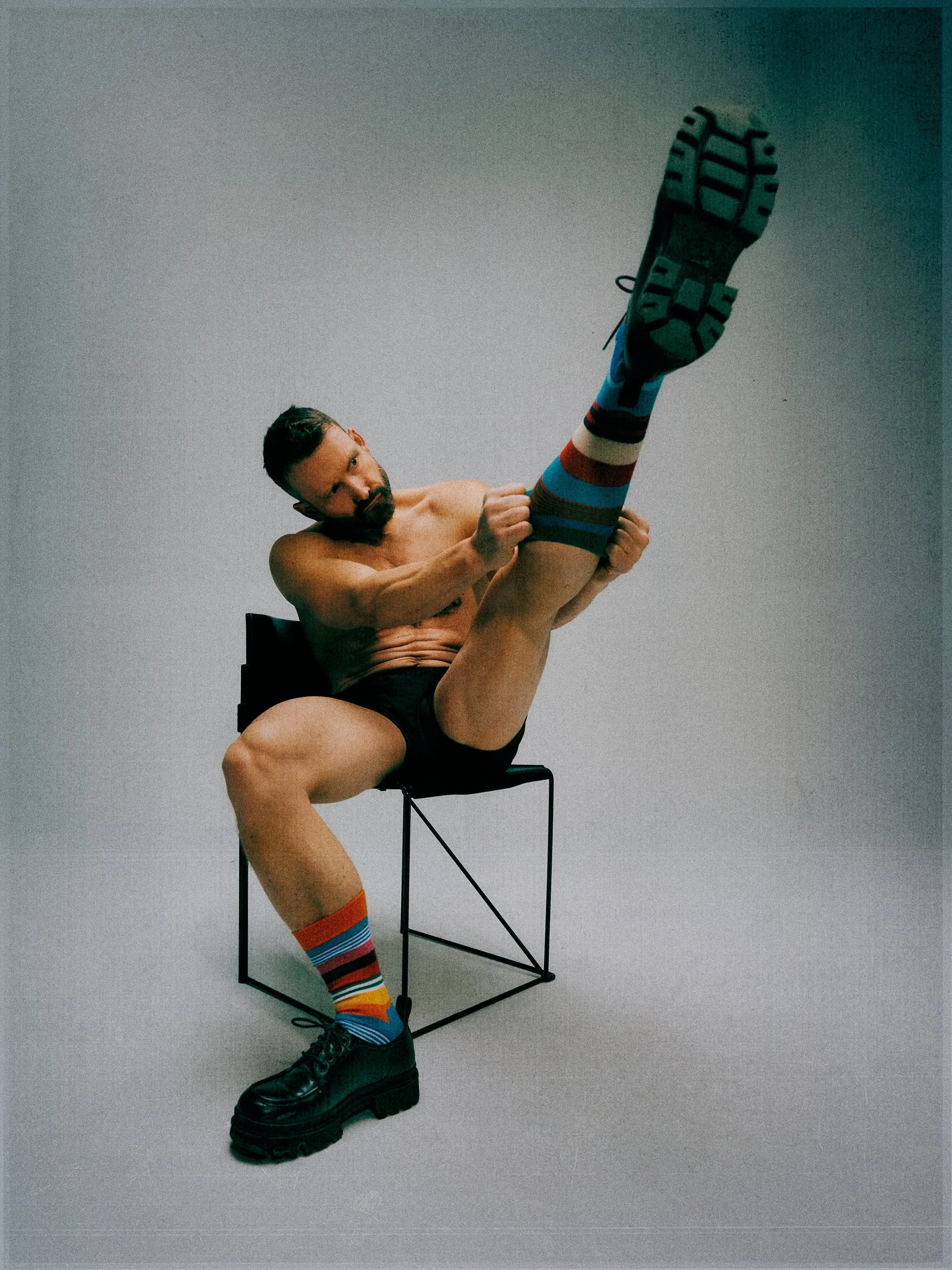 Shirtless man seated on a metal chair holding one raised leg, with the sole of the shoe facing the camera.