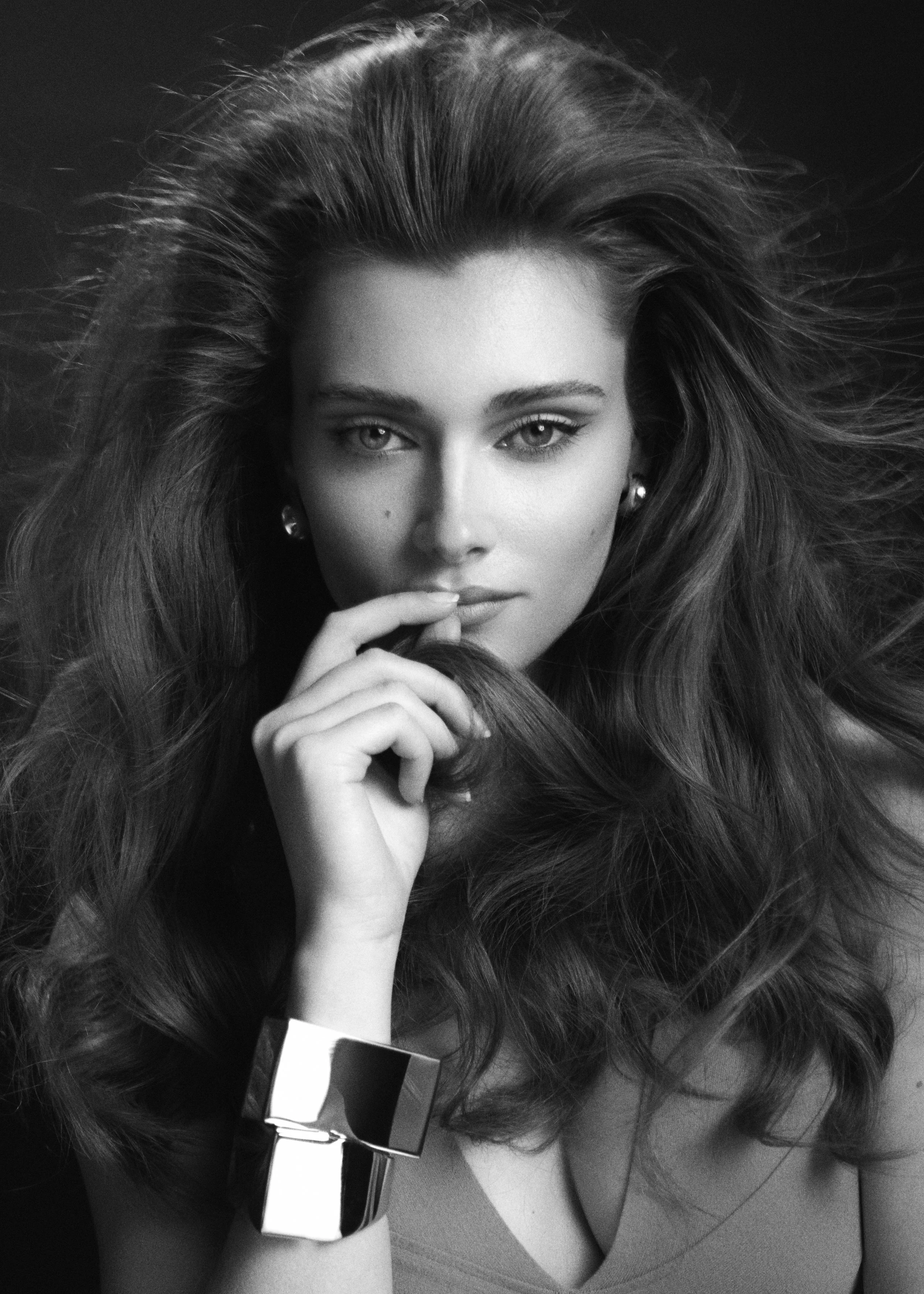 Black-and-white beauty portrait of a woman with voluminous hair and a finger resting beside her lips.