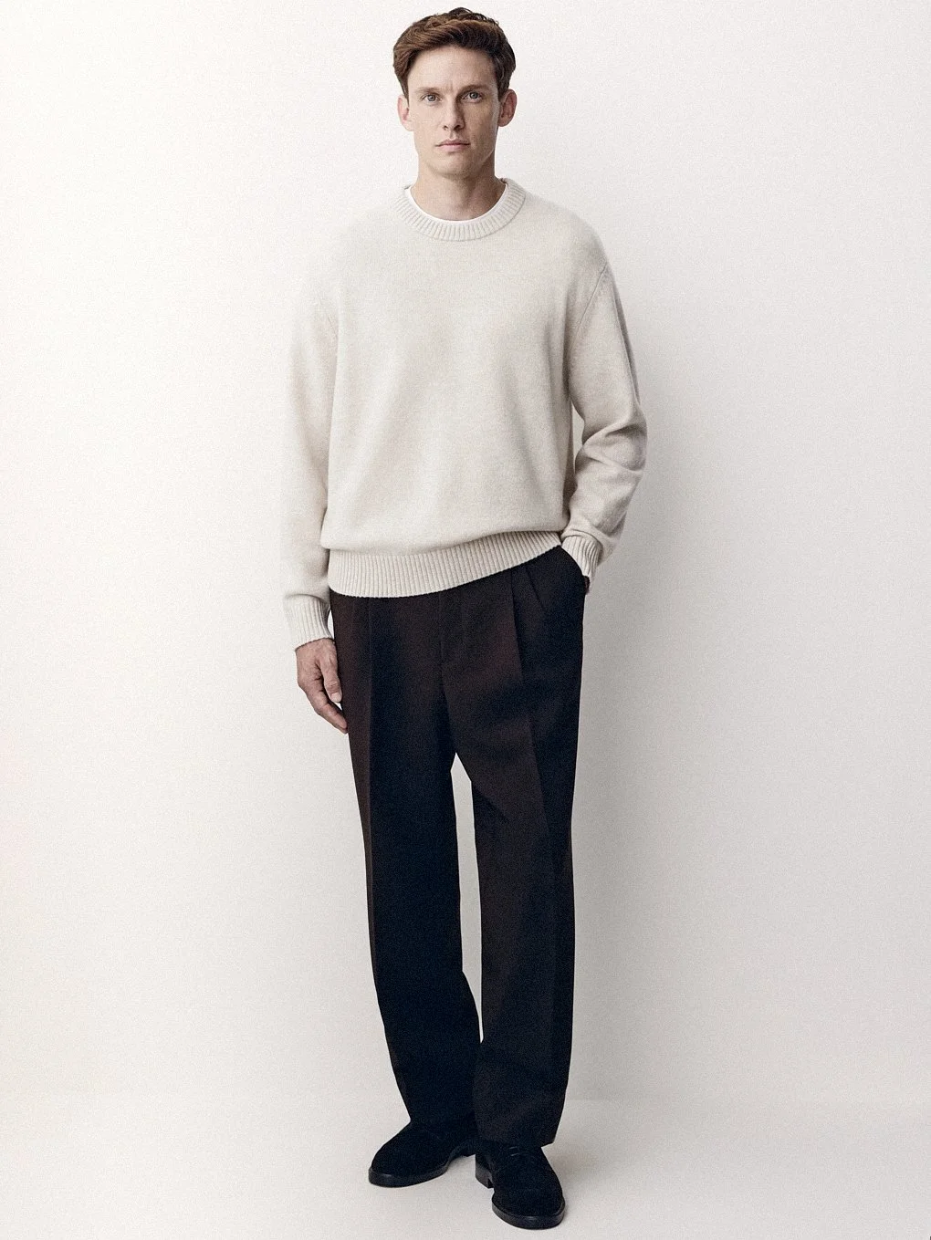 Man in a cream sweater and dark trousers standing against a pale studio wall.
