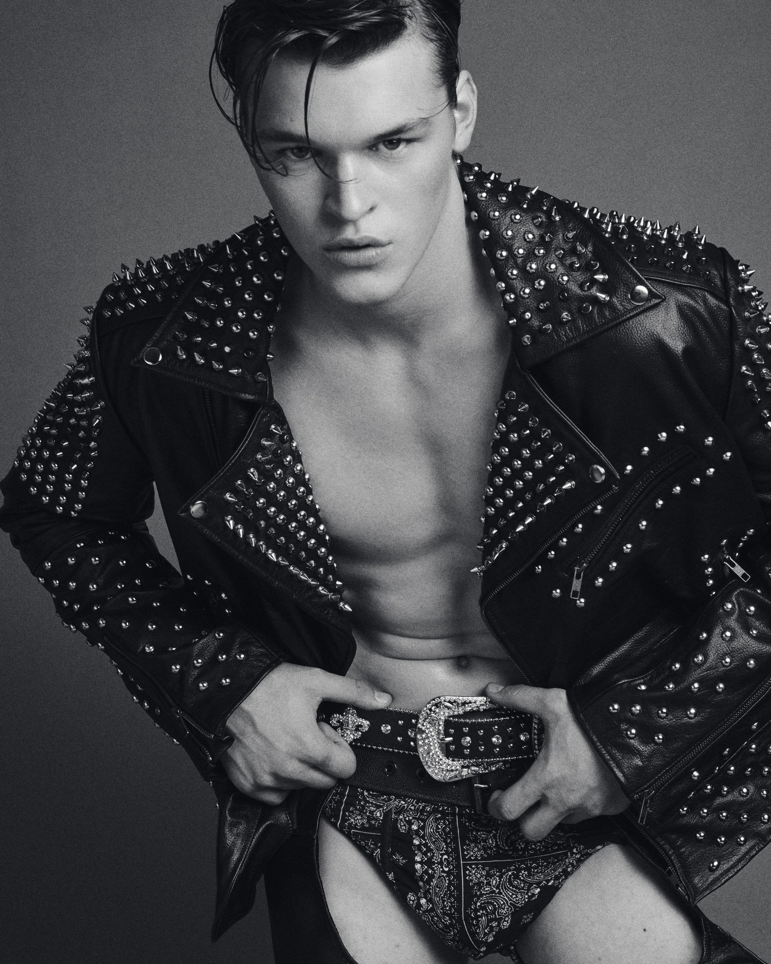 Black-and-white portrait of a shirtless man in a studded leather jacket with his hands at his belt.