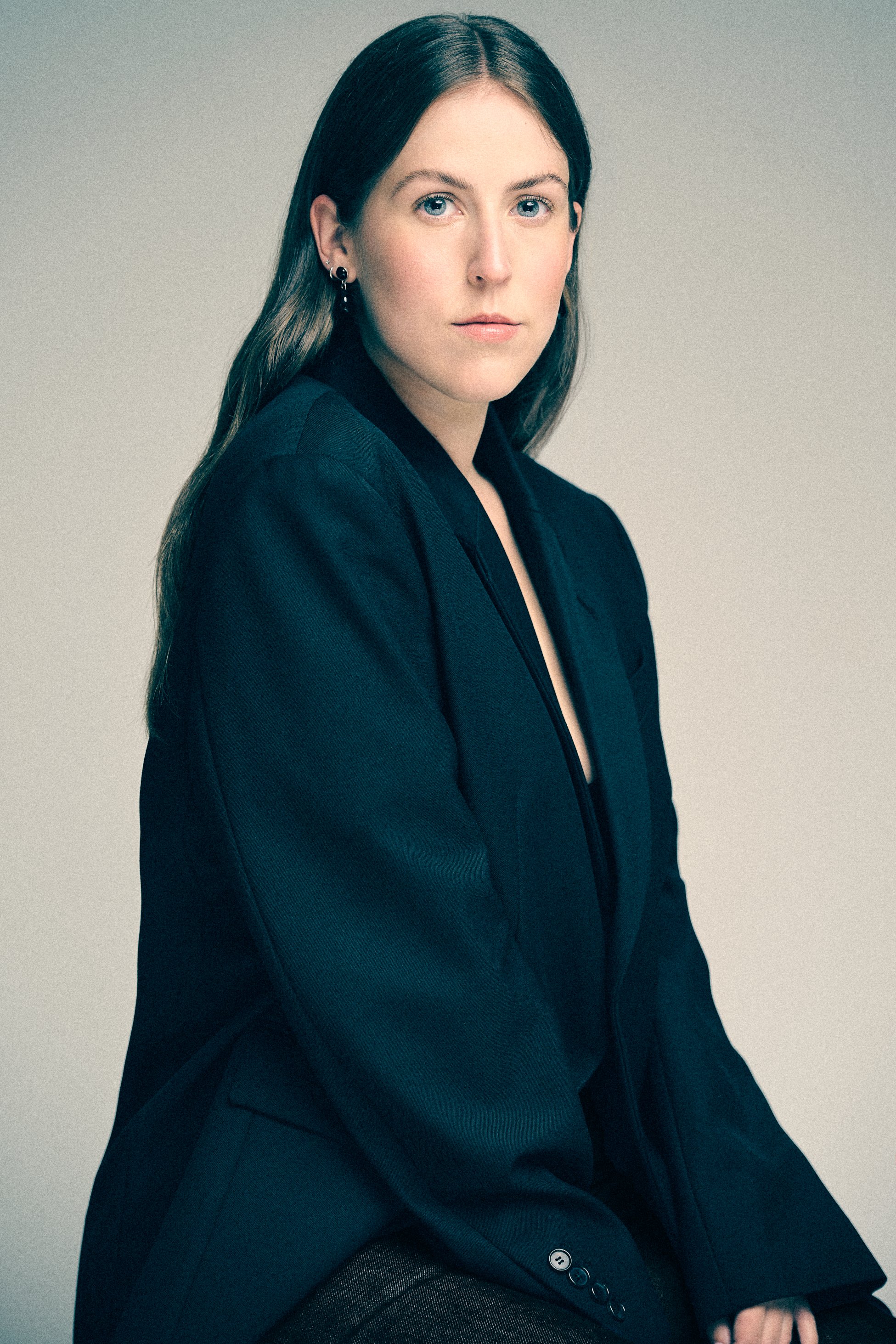 Woman with long dark hair seated in a black blazer against a pale studio background.