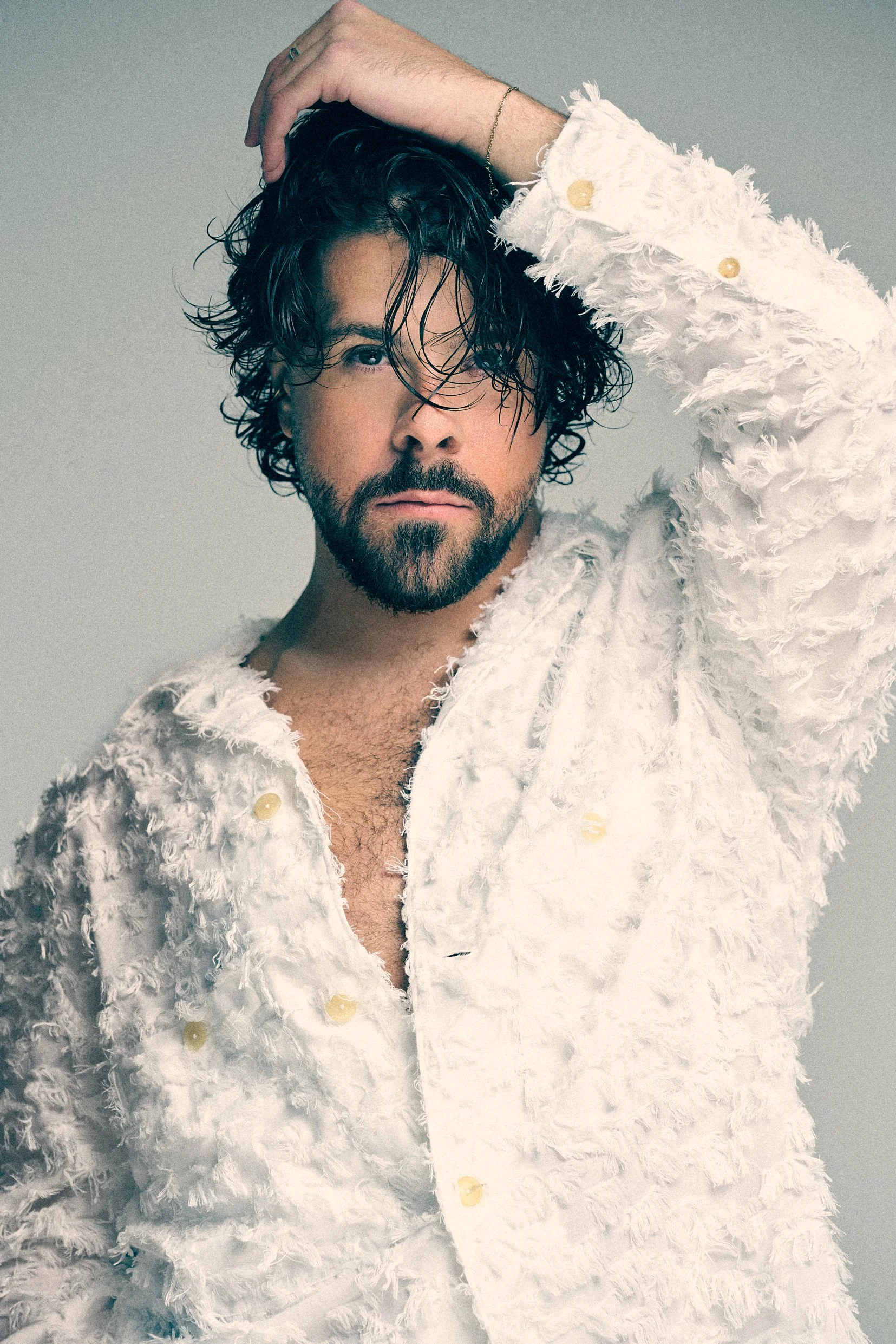 Bearded man with curly hair wearing a textured white shirt and lifting one hand above his head.