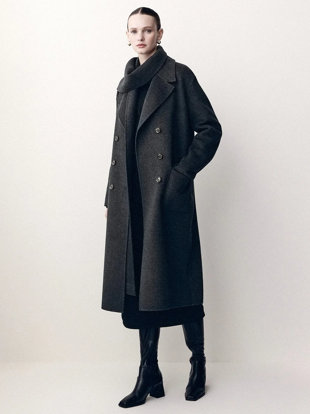 Woman in a long dark coat and black boots, with her hands in the coat pockets.
