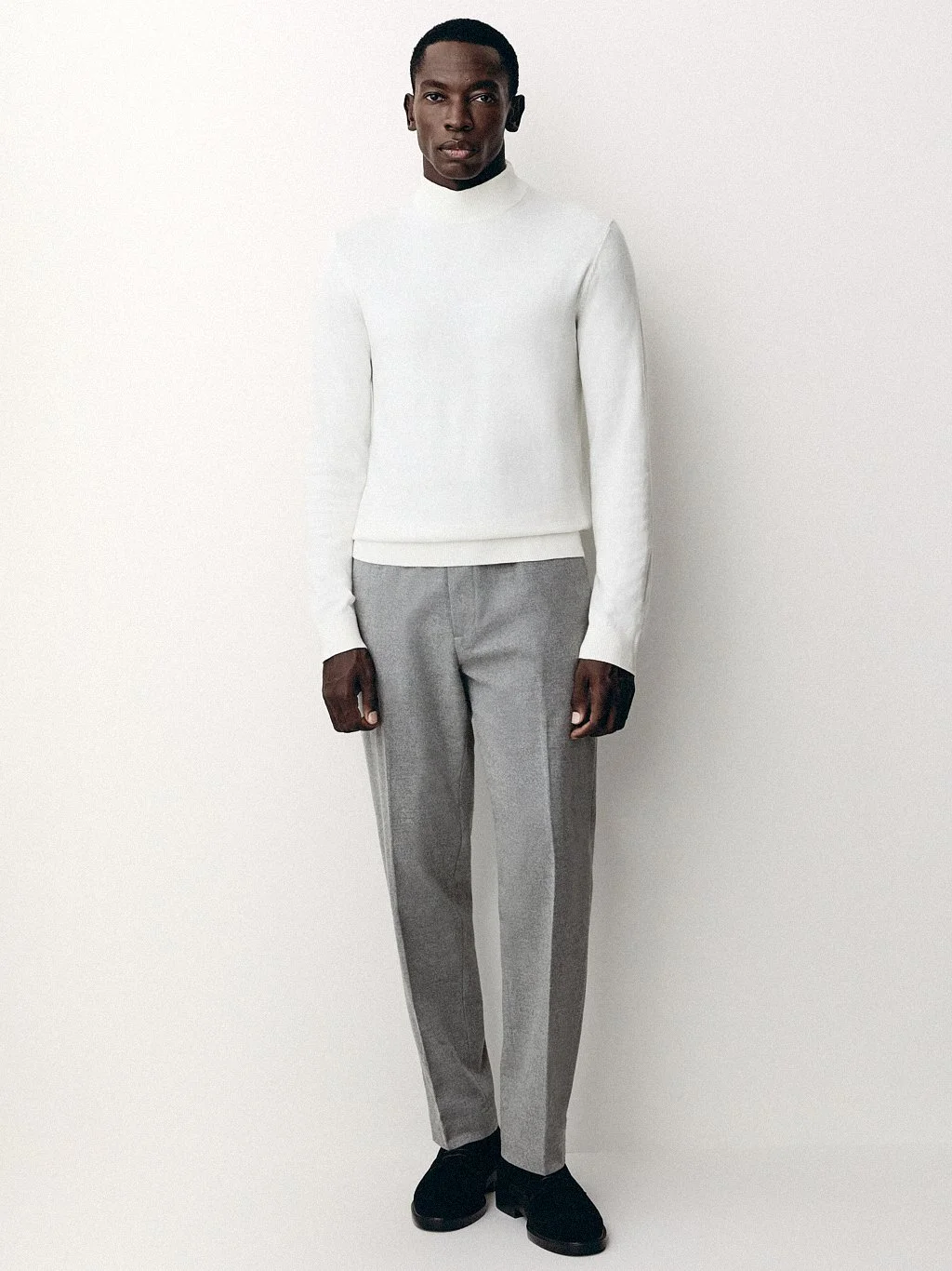 Man in a white sweater and grey trousers standing against an off-white studio background.
