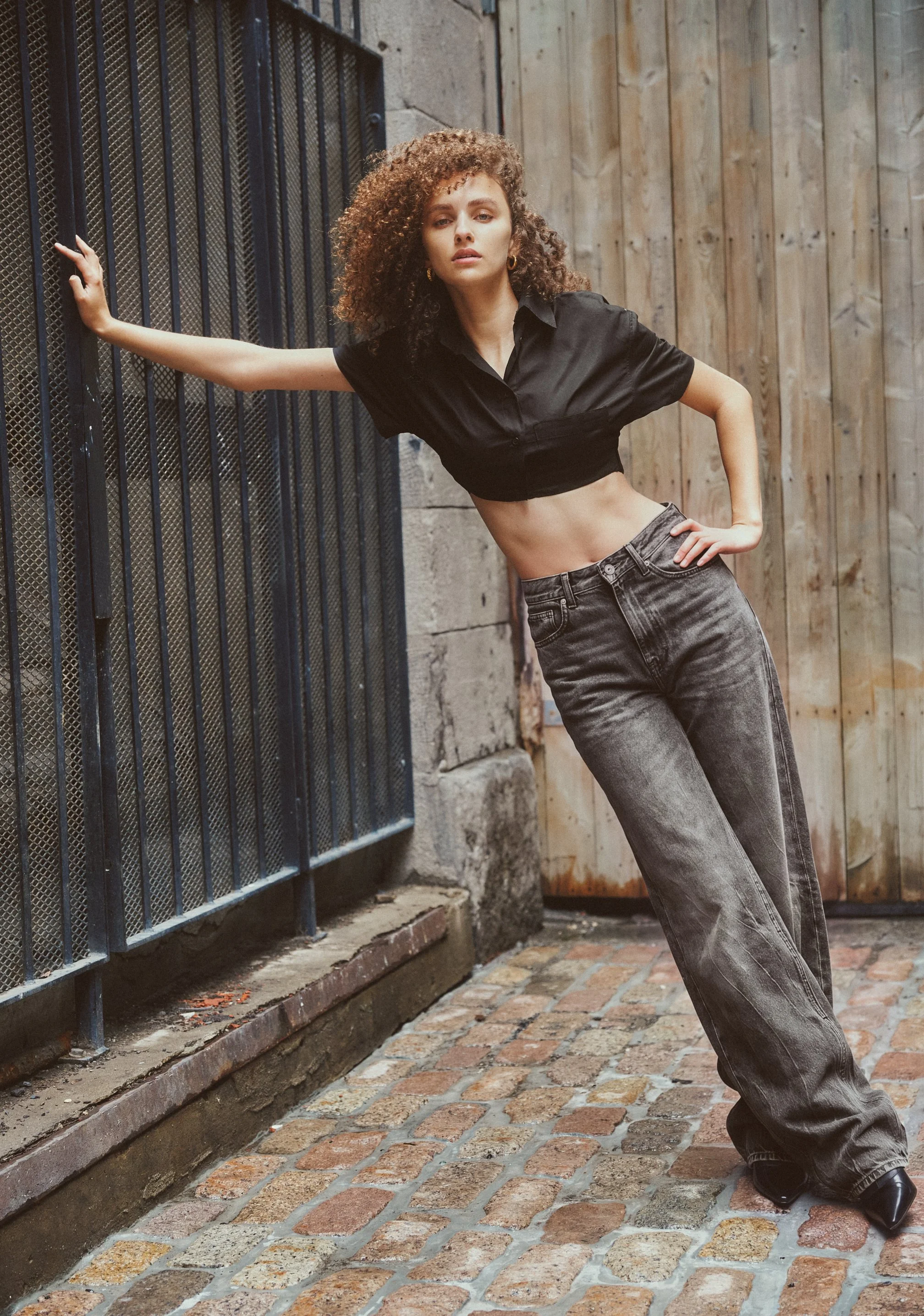 Full-length fashion photograph of a model in a cropped black shirt and wide-leg grey jeans leaning sideways against an iron gate.