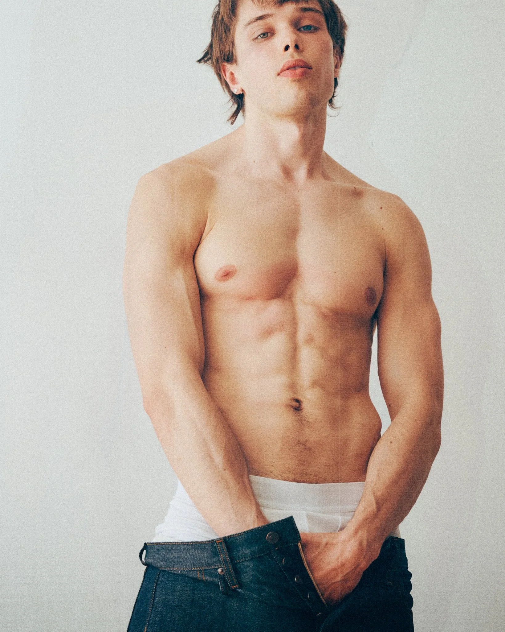 Shirtless man looking at the camera with his hands holding the waistband of dark jeans.