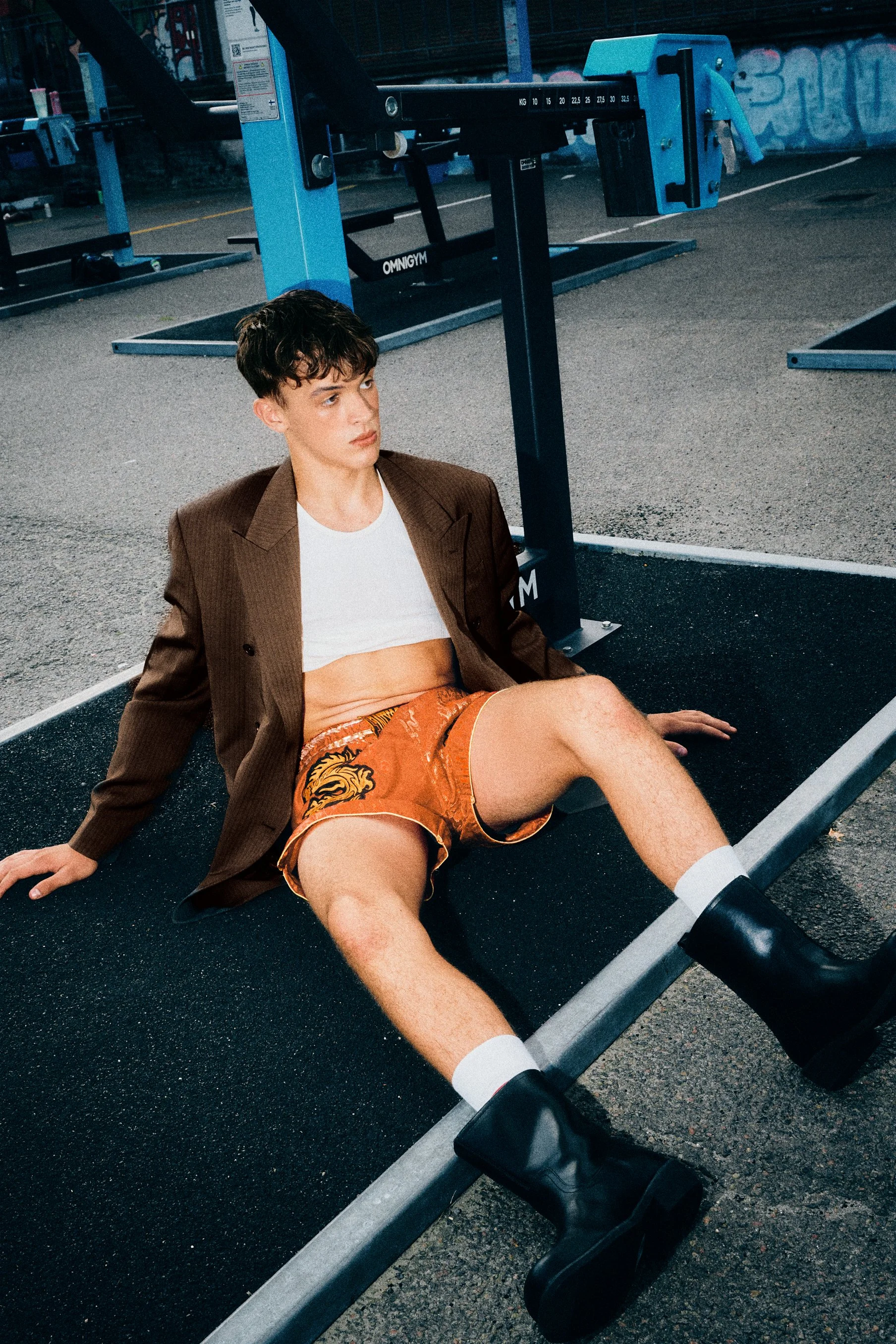 Man seated in a brown blazer, cropped white top and orange shorts.