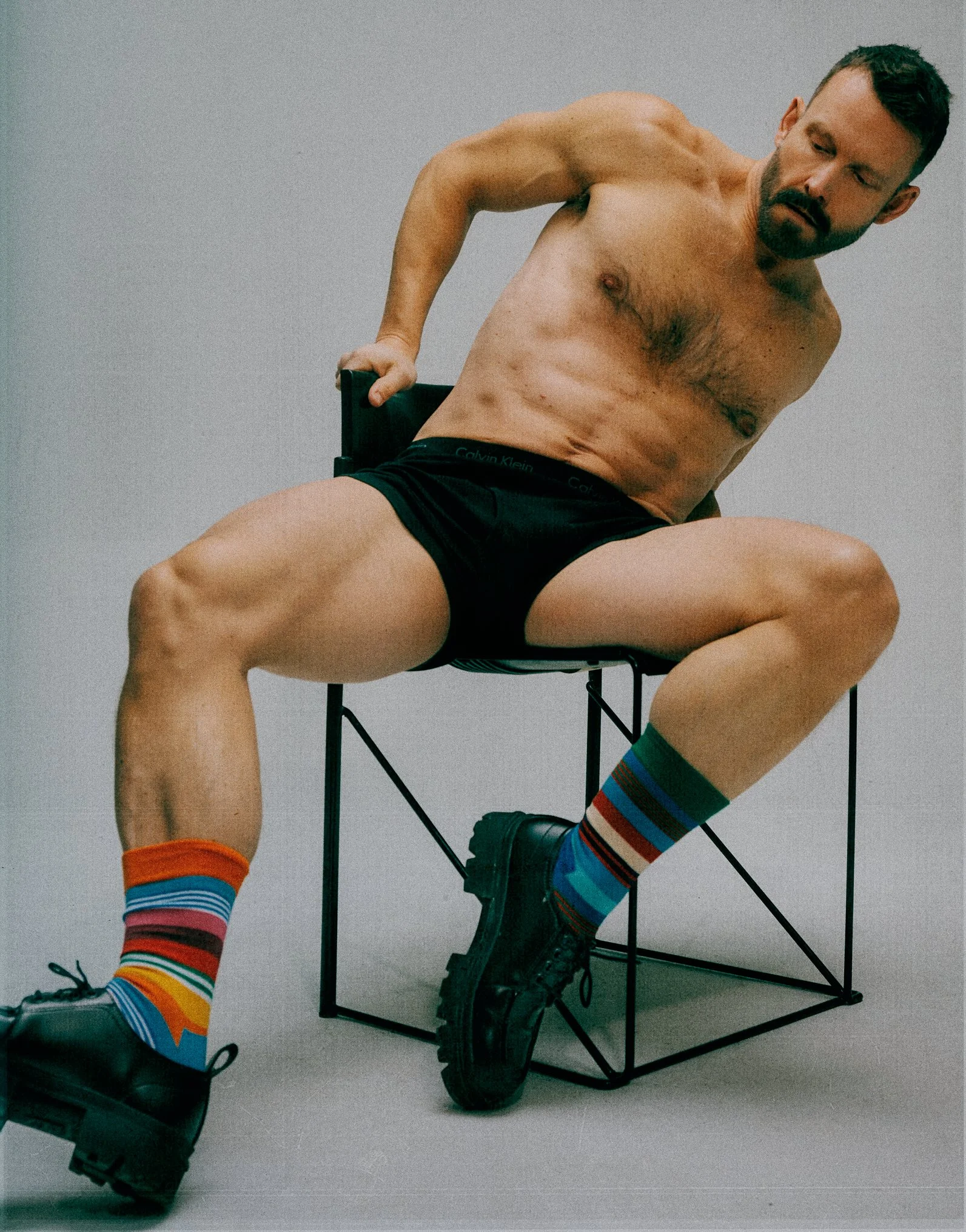 Shirtless man sitting with his legs apart on a metal chair in black shorts and colourful striped socks.
