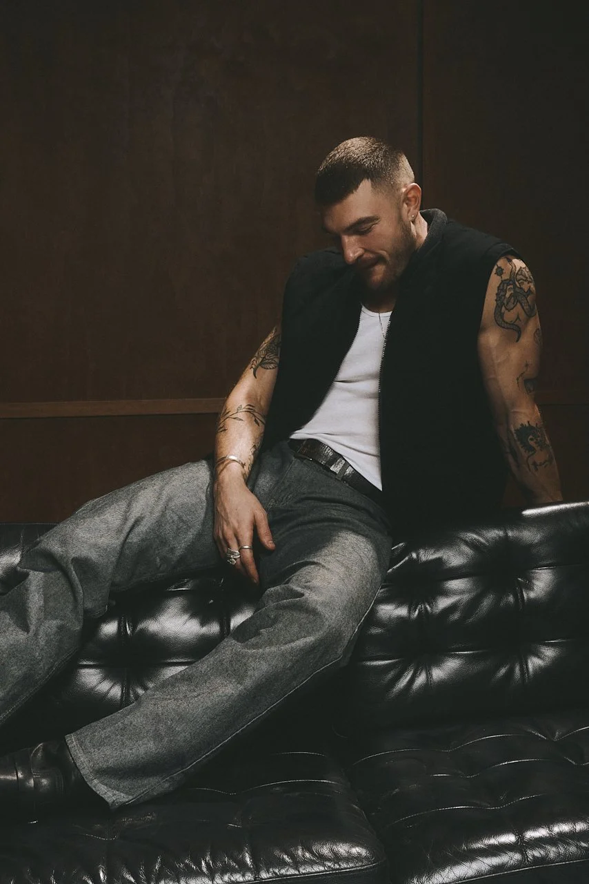 Tattooed man in a dark sleeveless vest and jeans sitting loosely on a black leather sofa.