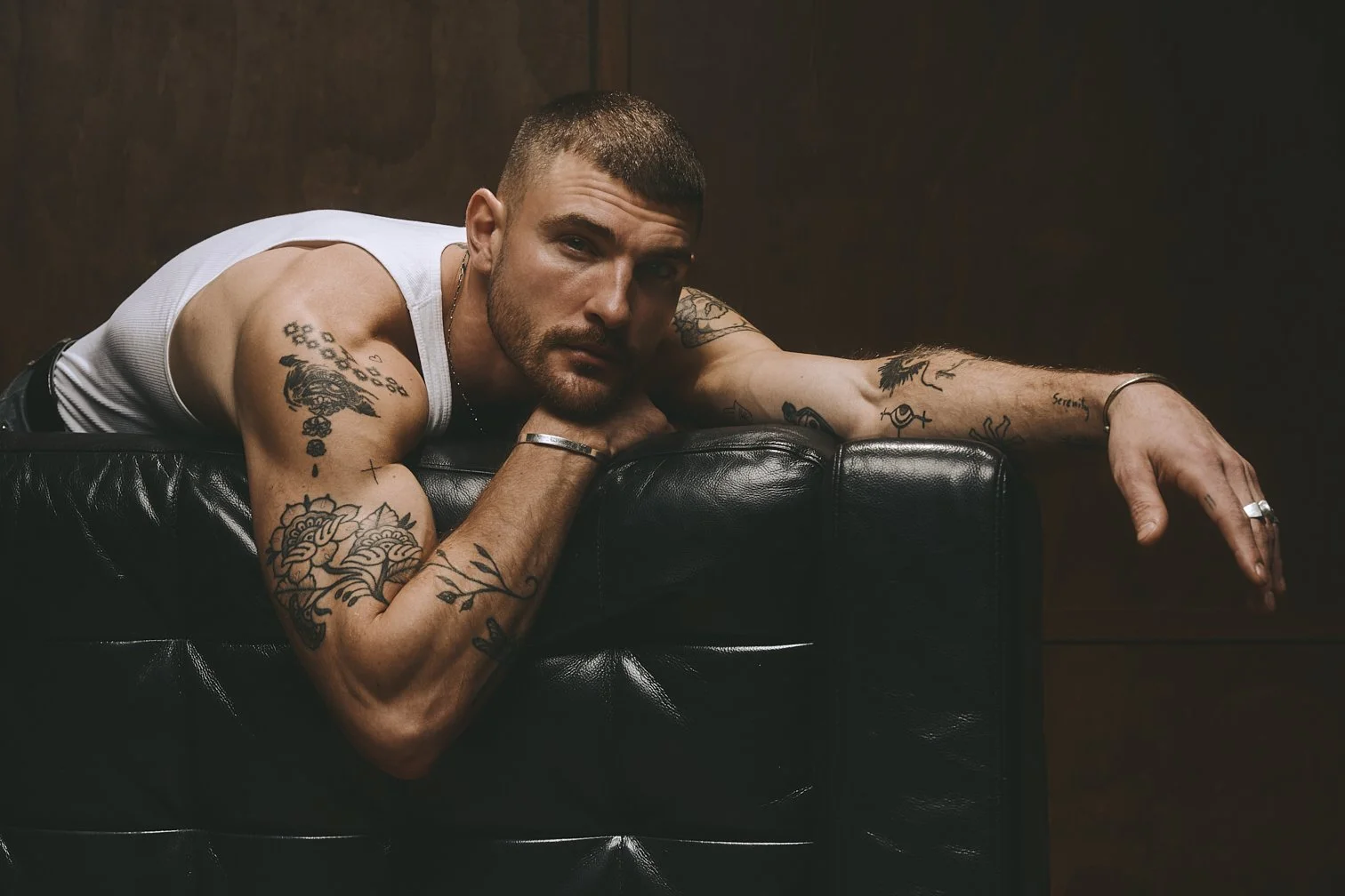 Tattooed man in a white tank top leaning over a leather sofa arm with his head resting on his hand.