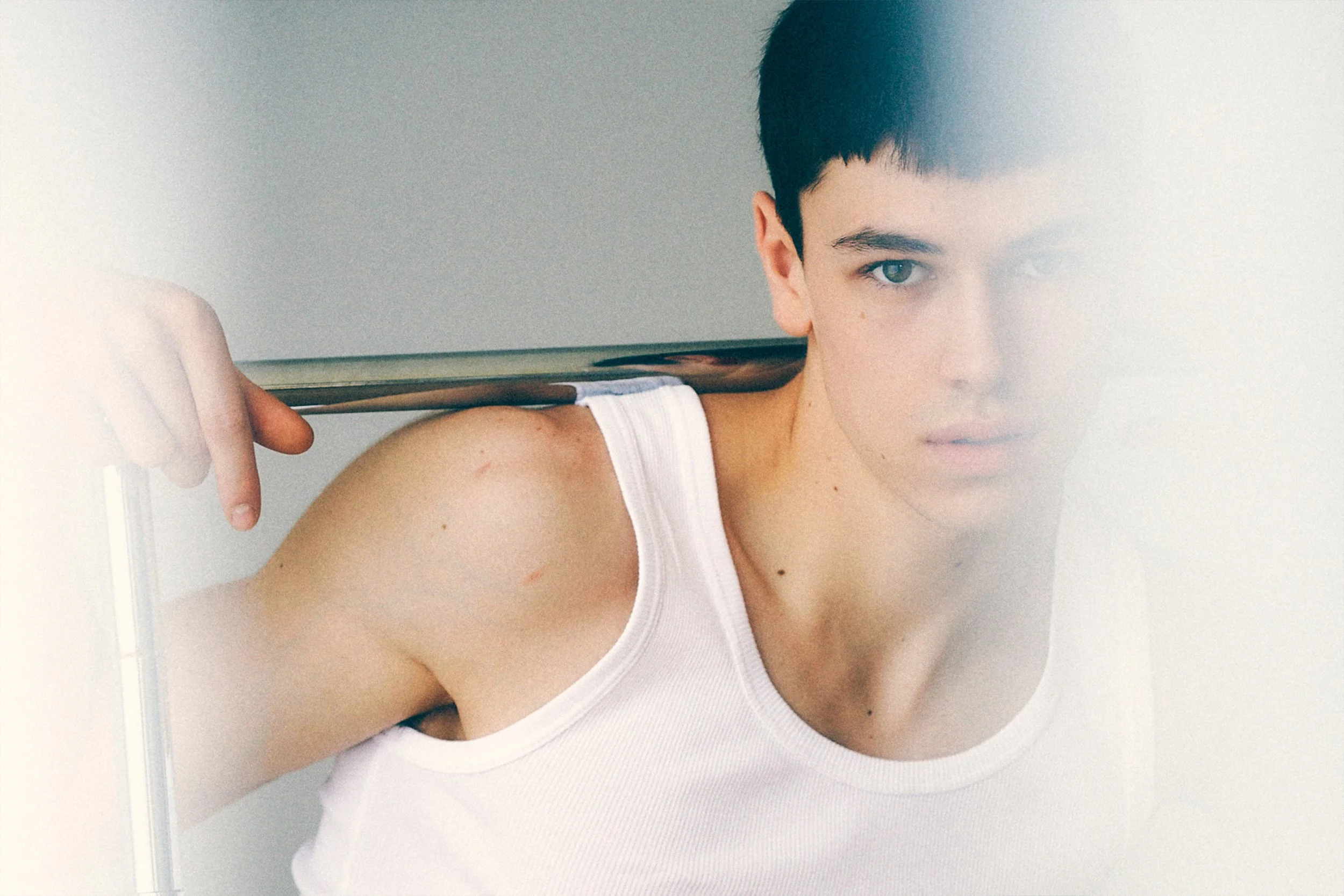 Close portrait of a young man in a white tank top partly obscured by a sheer curtain.