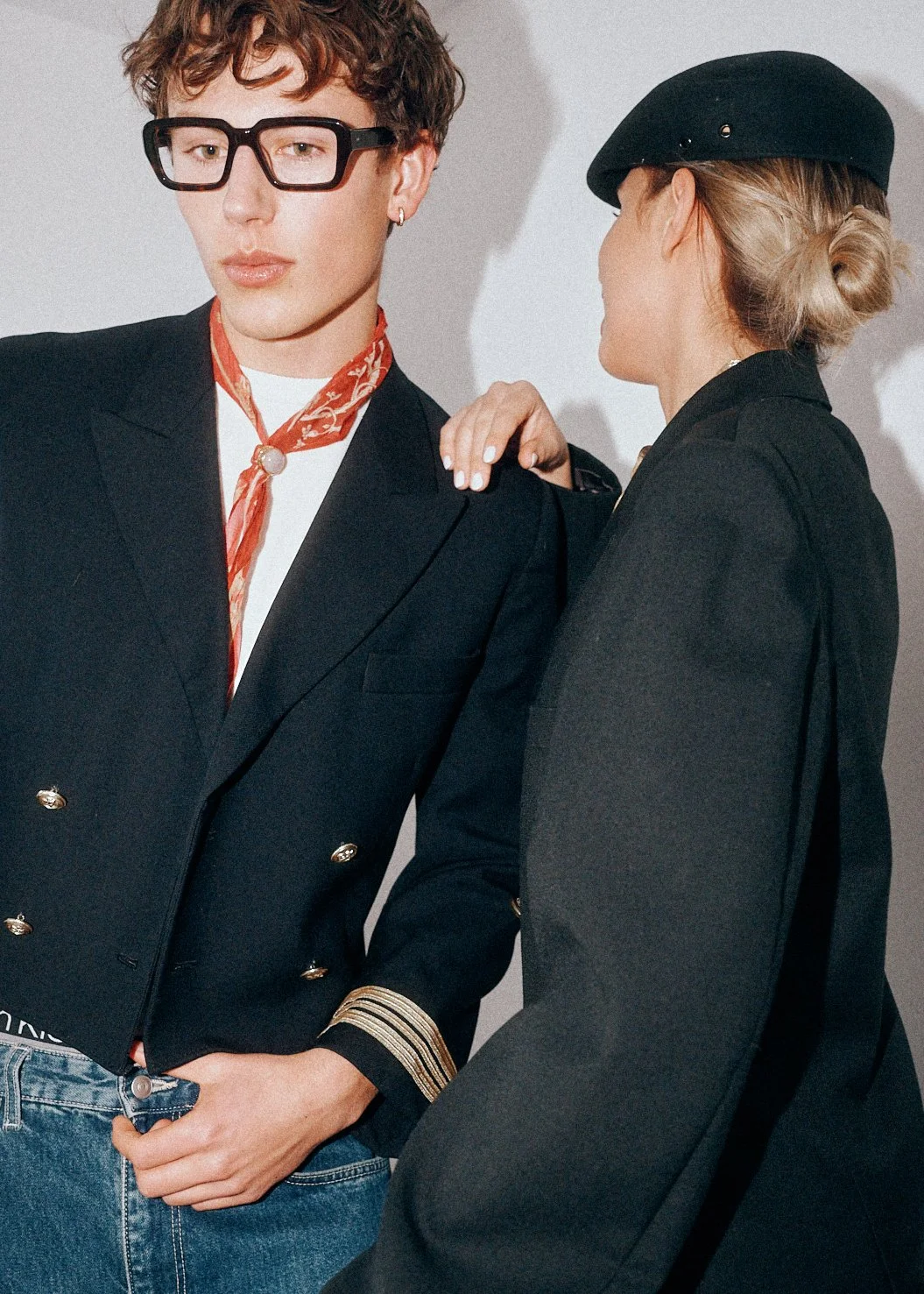 Two models in dark tailoring, one wearing oversized glasses and the other a black beret.
