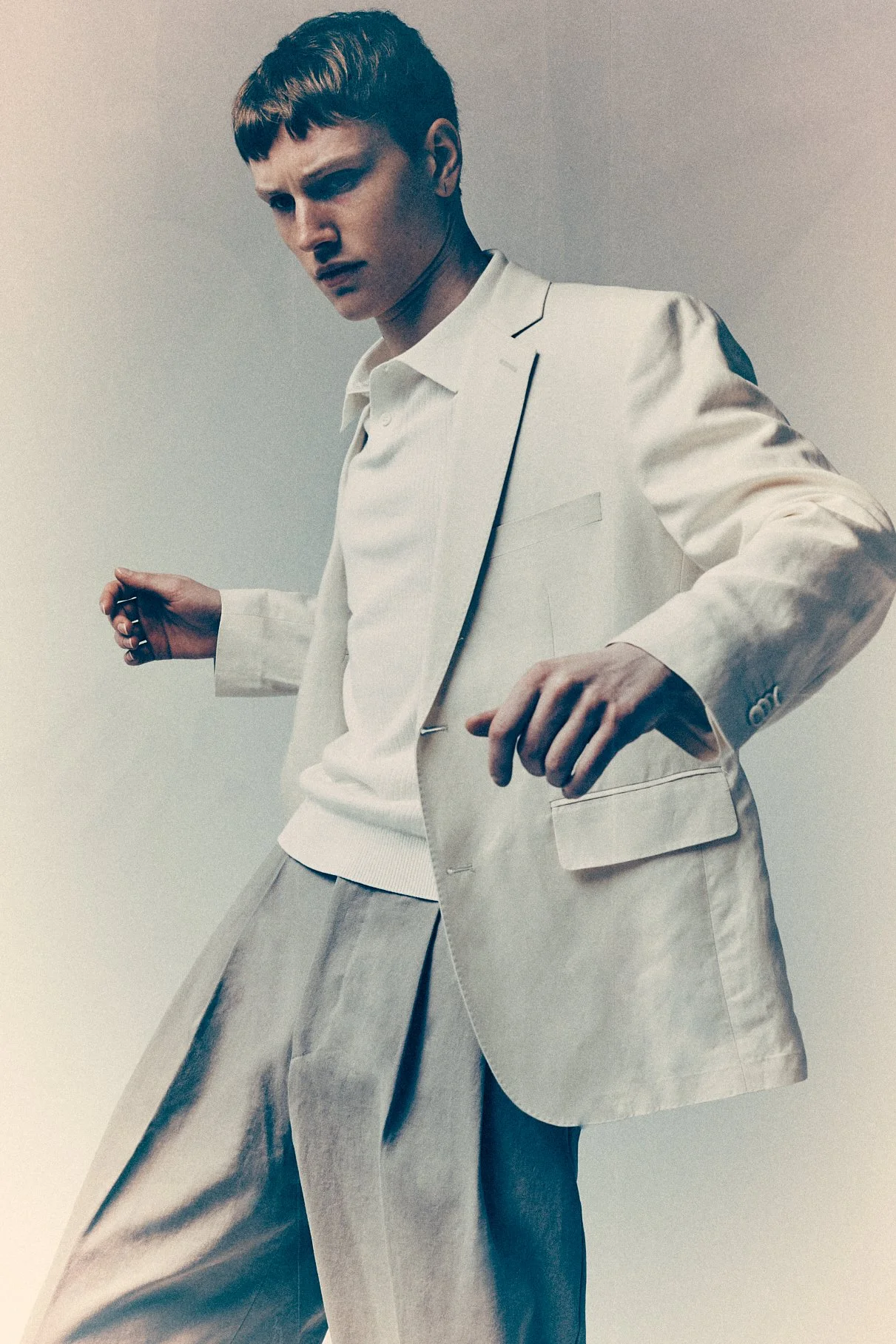 Man leaning forward in a cream blazer and pale shirt with both arms lifted slightly.