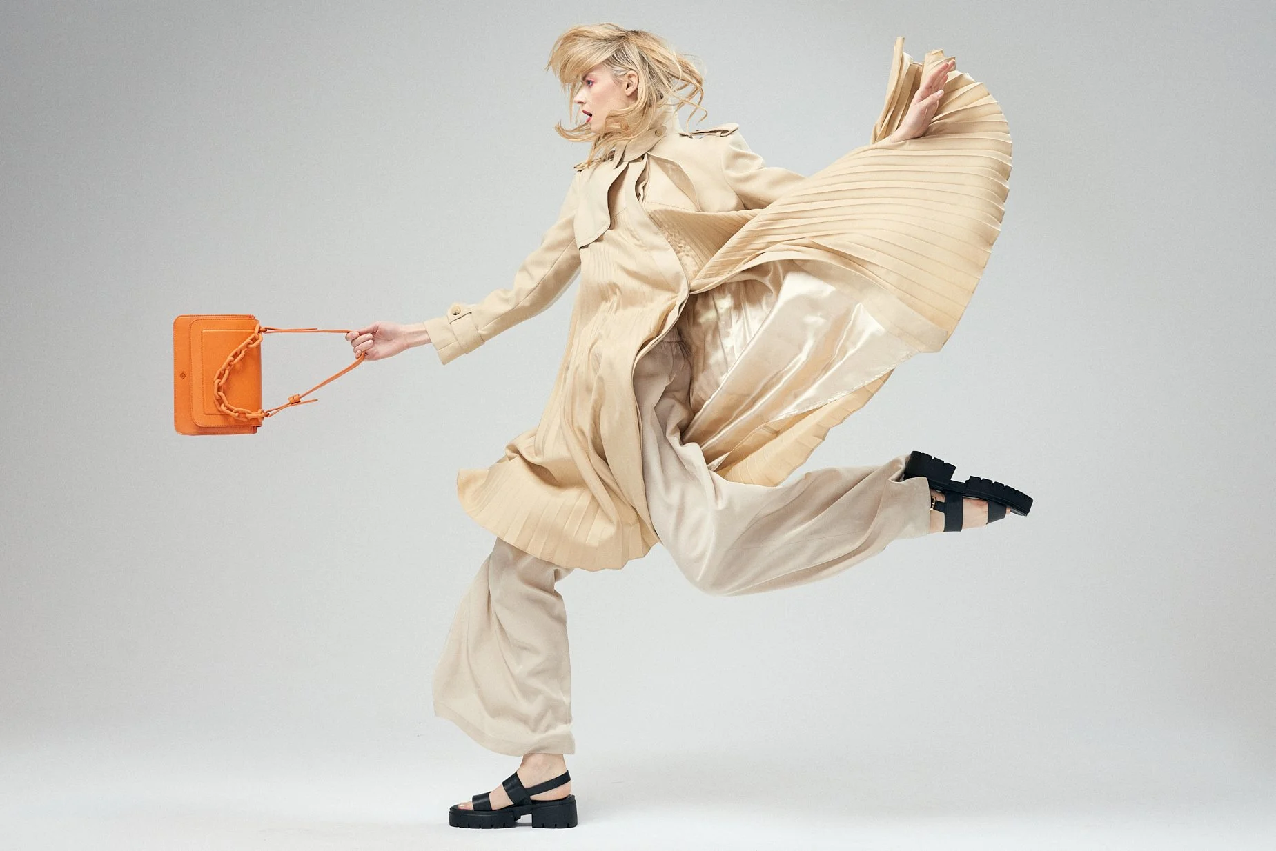 Blonde woman leaping in a flowing beige coat while holding a small orange handbag.