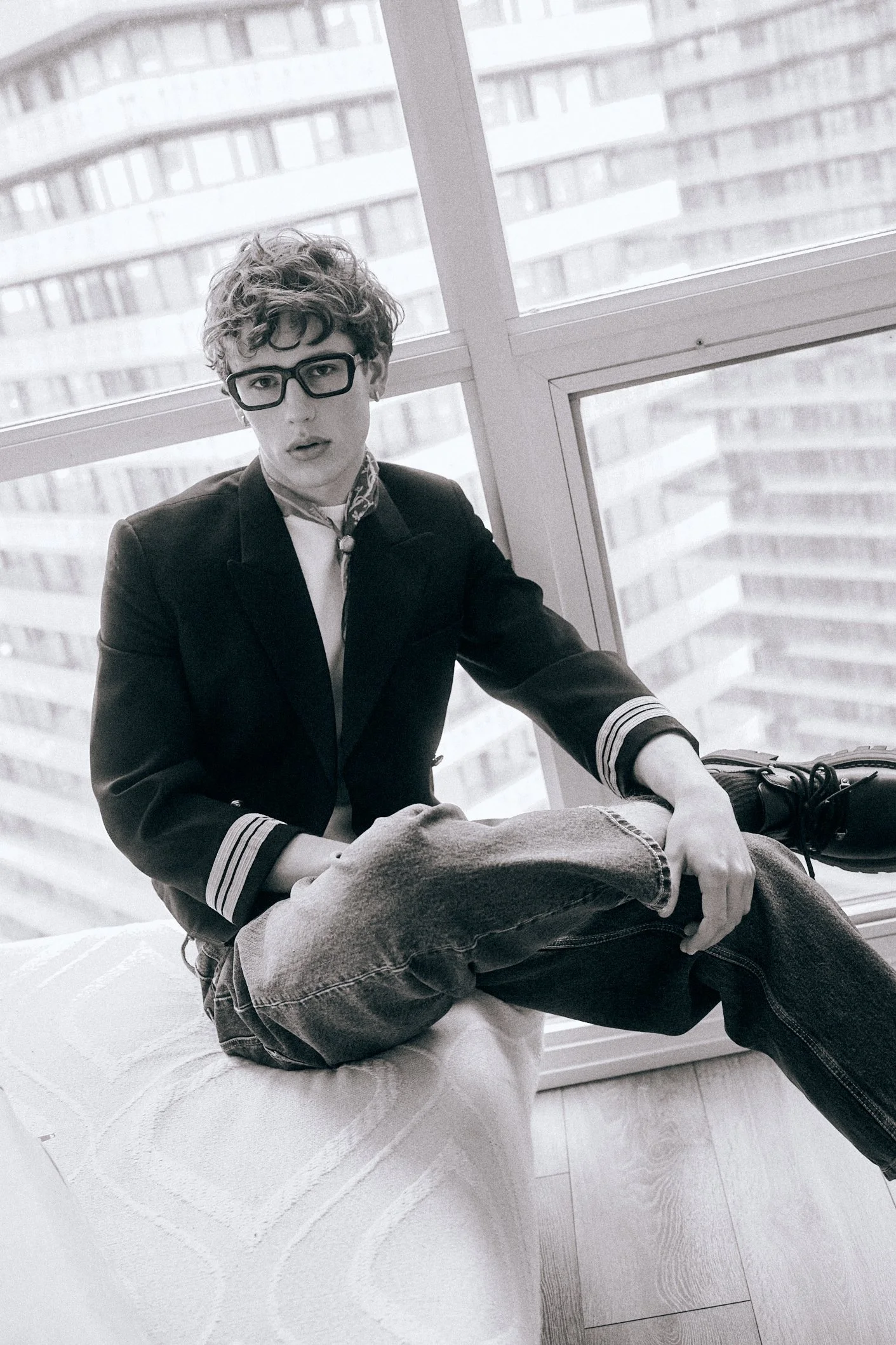 Black-and-white portrait of a man in glasses and a black blazer sitting beside a large window.