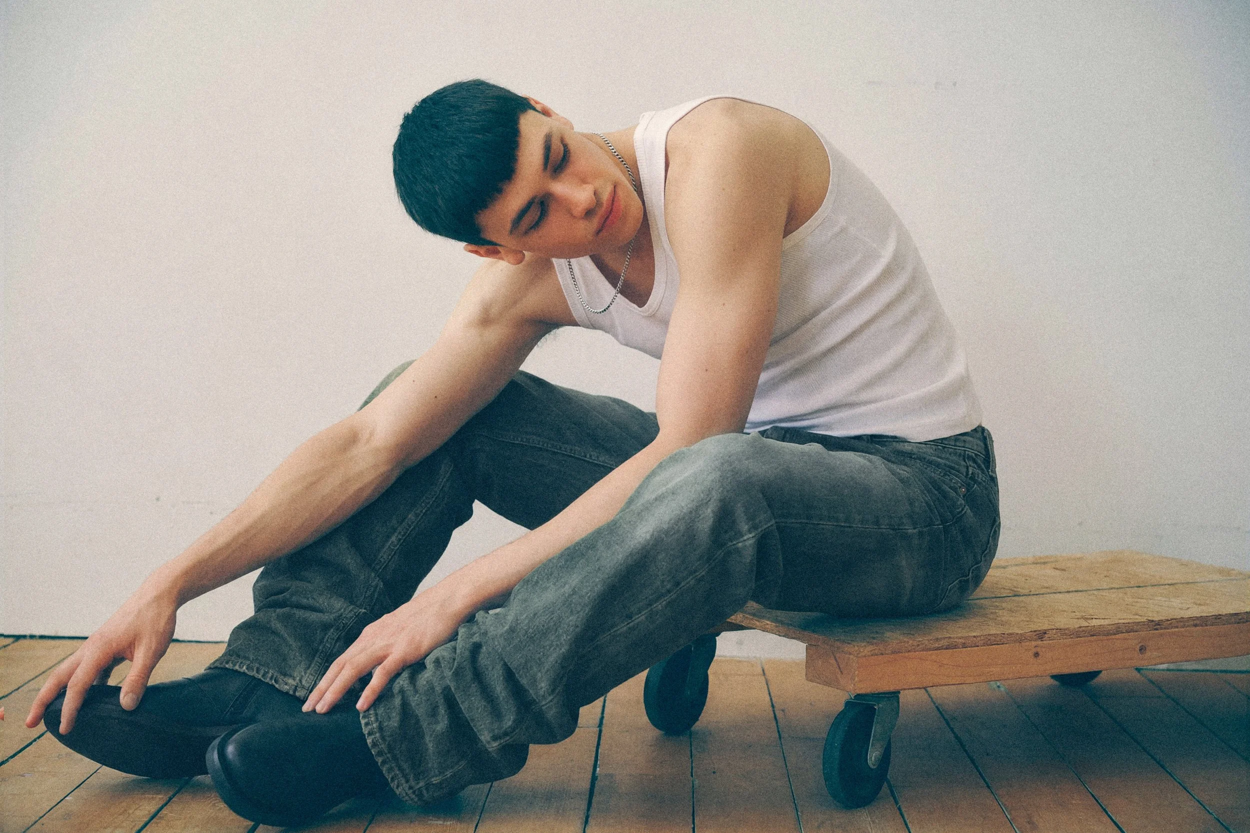 Young man in a white tank top and jeans seated sideways on a low wooden bench.