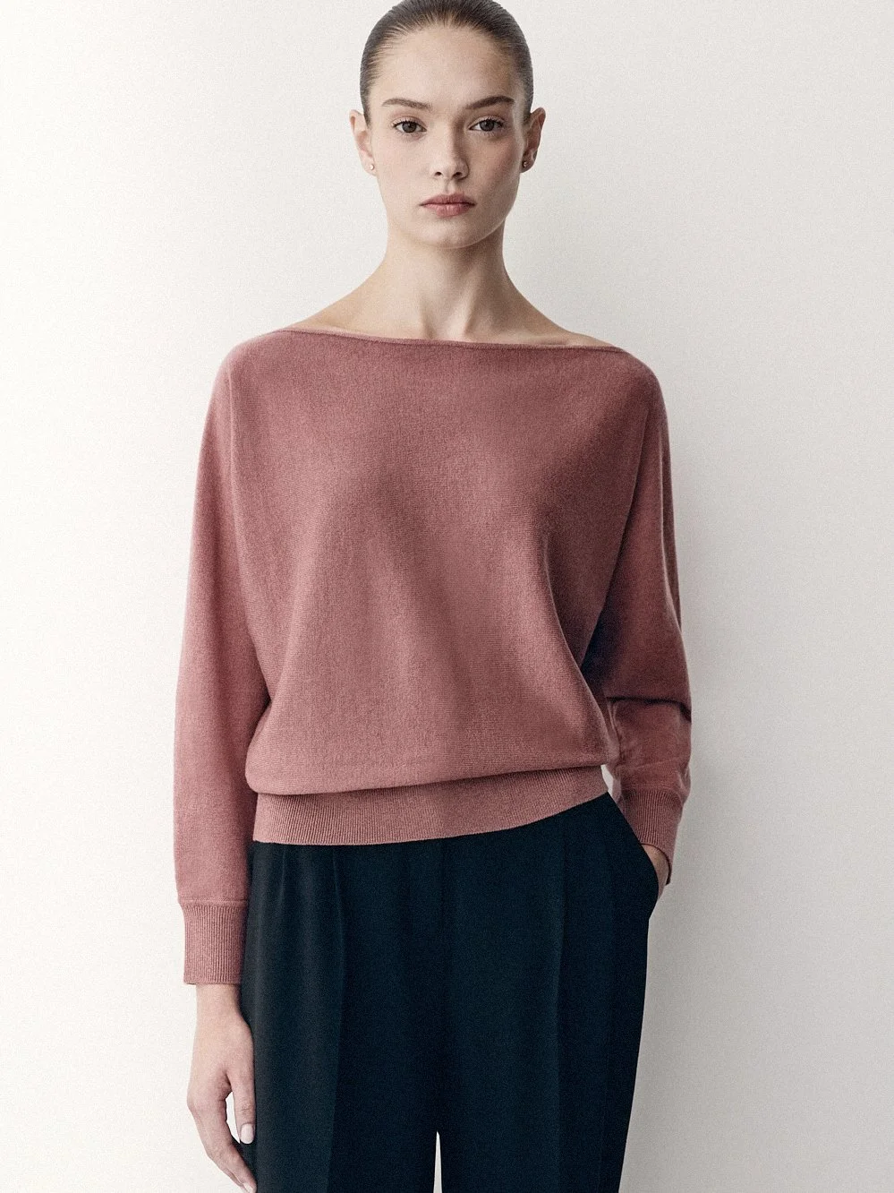 Woman in a dusty pink sweater and dark trousers, with one hand in her pocket.