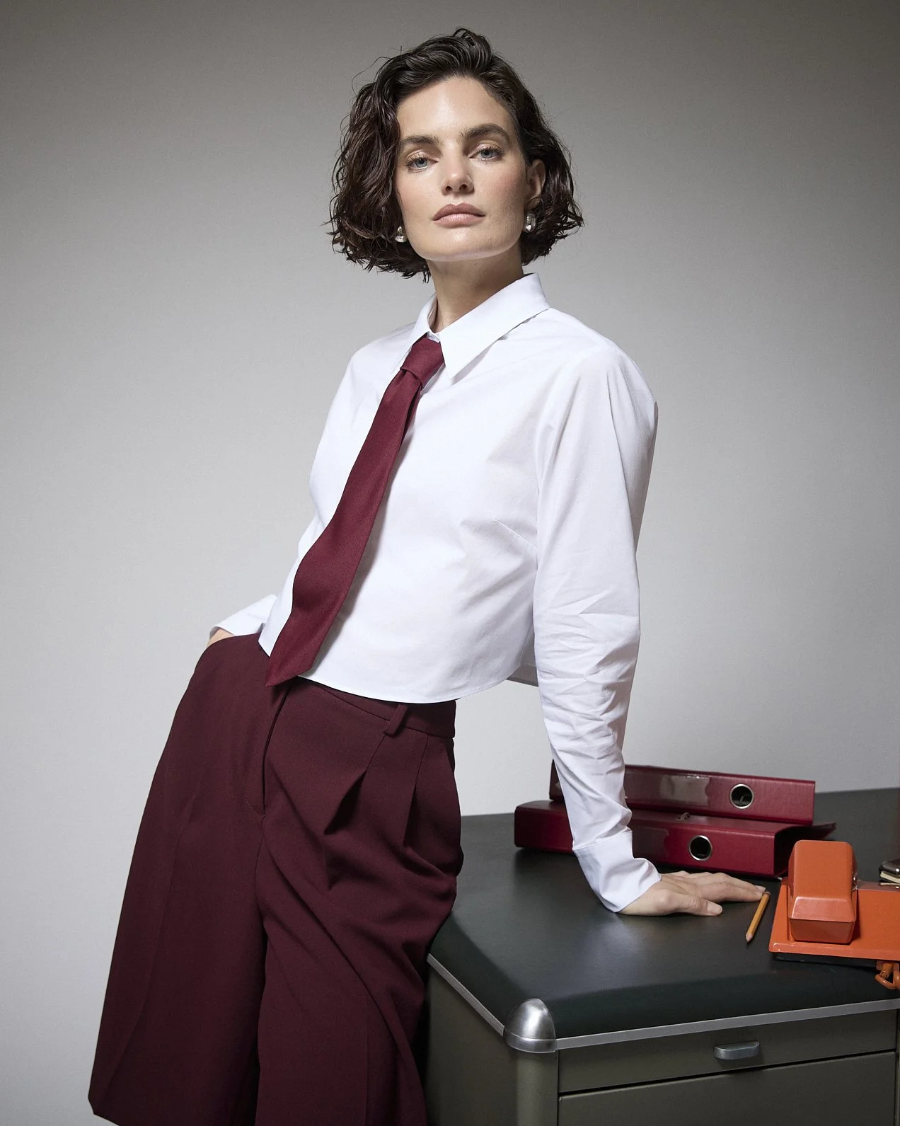 Woman in a white shirt, burgundy tie and trousers leaning against an office desk.