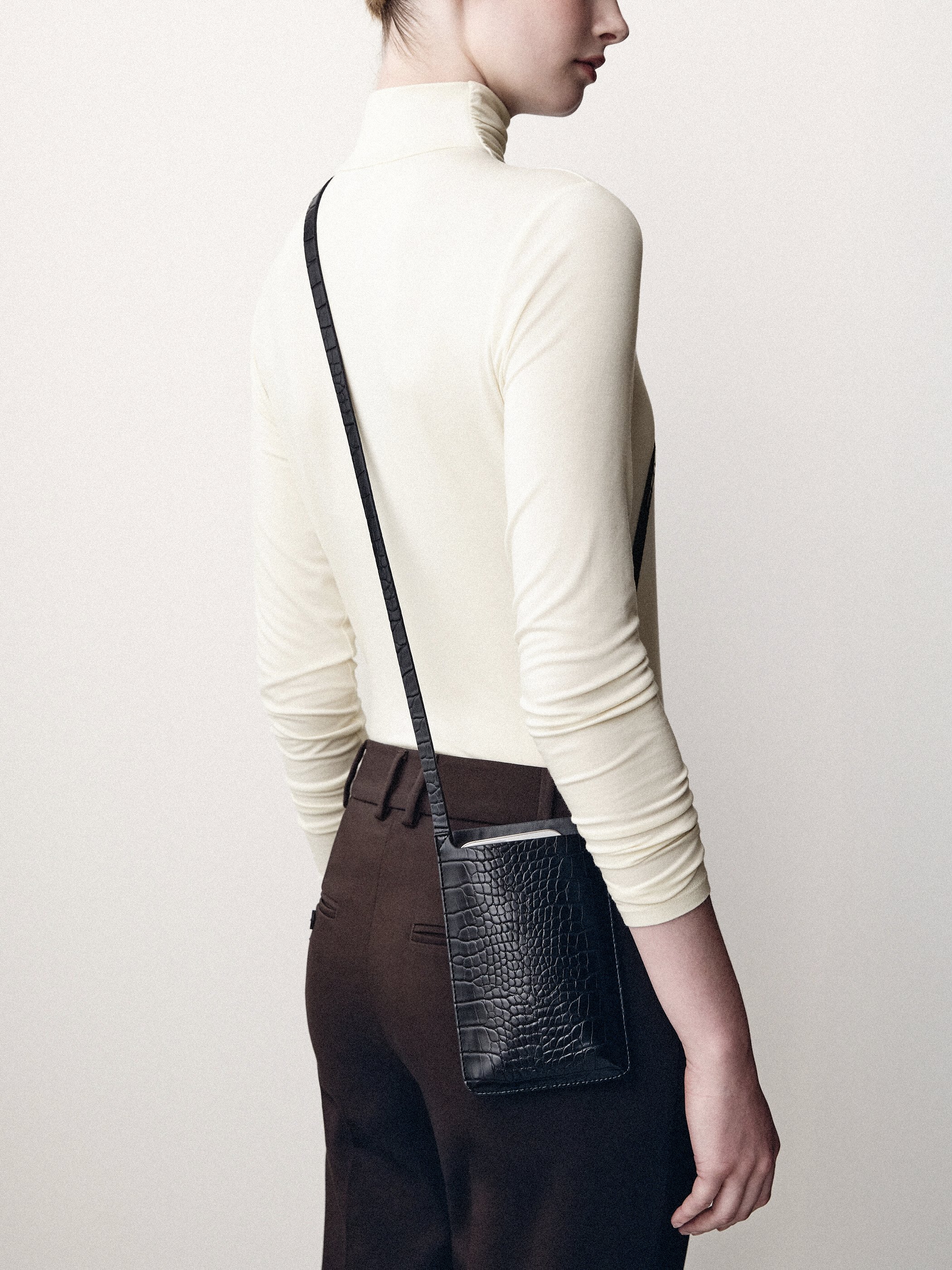 Back view of a cream turtleneck and dark trousers with a small black shoulder bag.