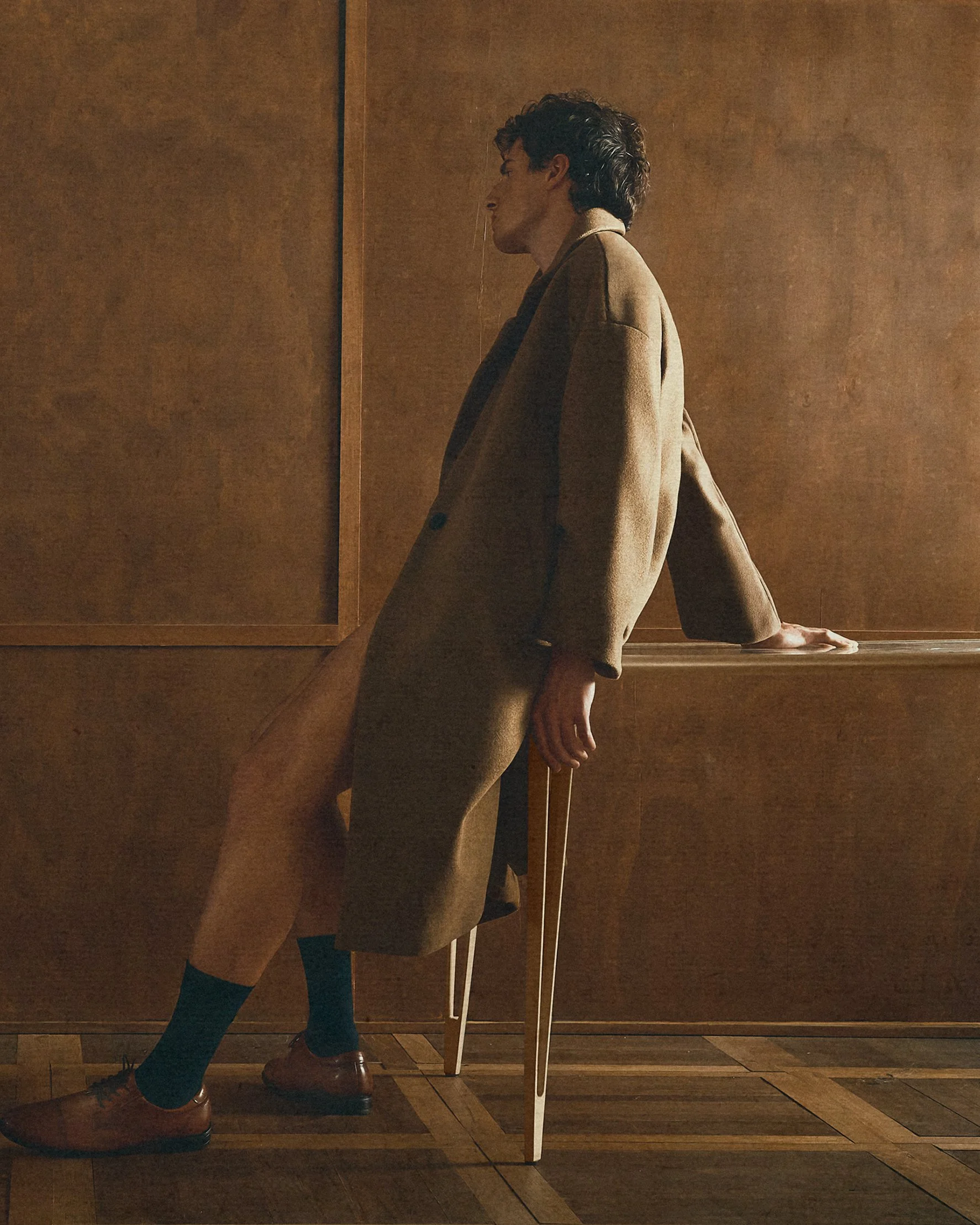 Side view of a man leaning back against a table in a beige coat and trousers.