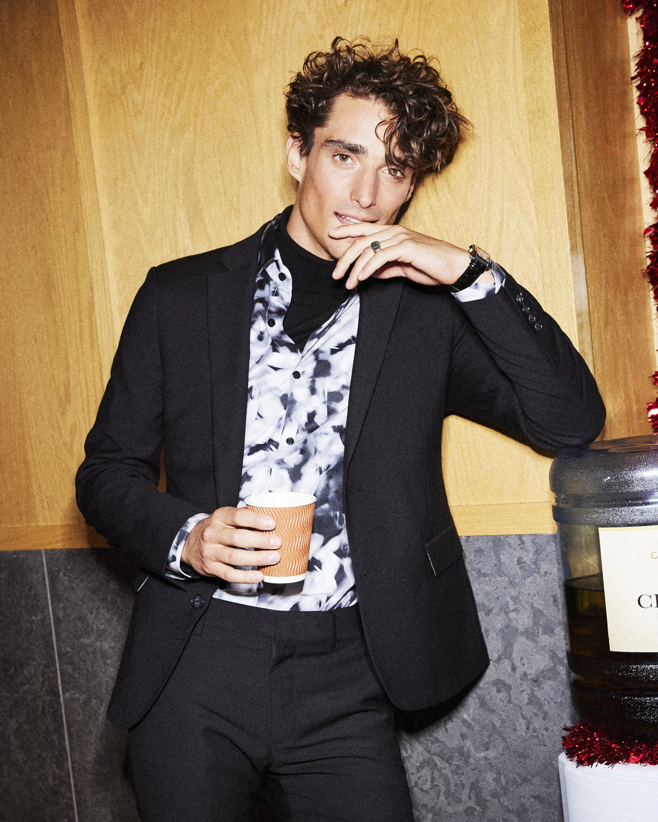 Man in a black suit and patterned shirt holding a paper cup beside a water cooler.