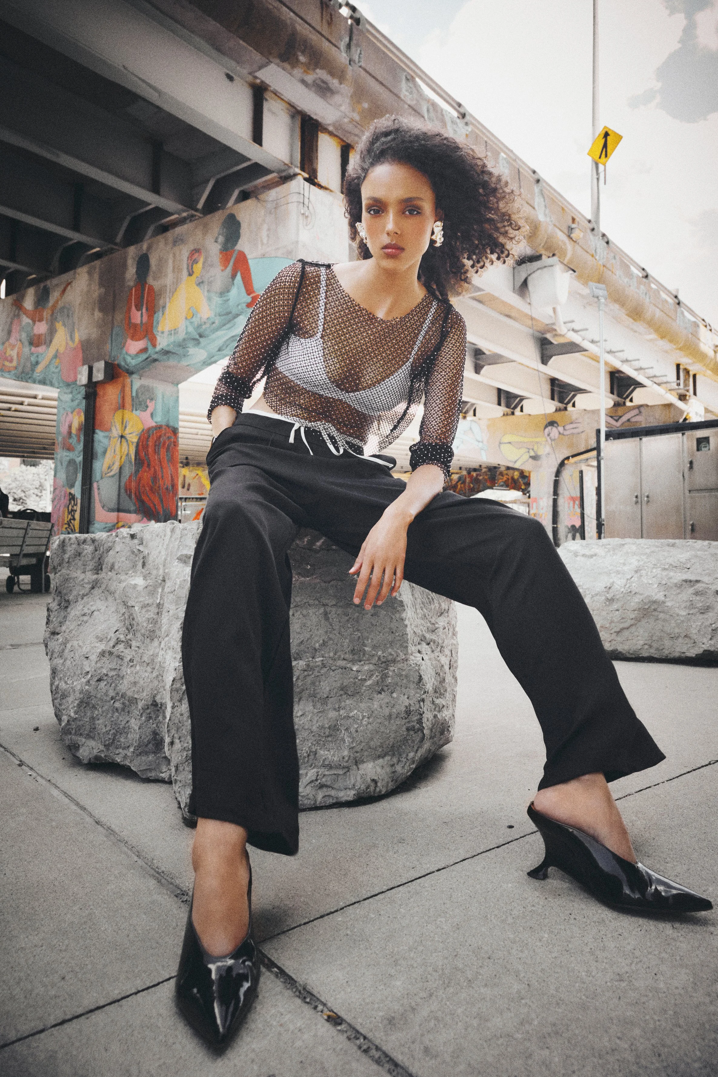 Woman in a sheer black top and cropped trousers seated on a concrete block.