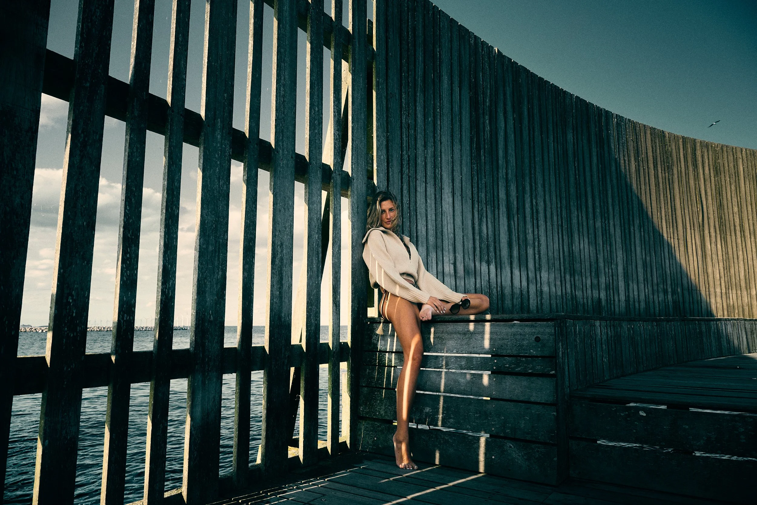 Woman leaning against a wooden bench inside a sunlit slatted waterfront structure.