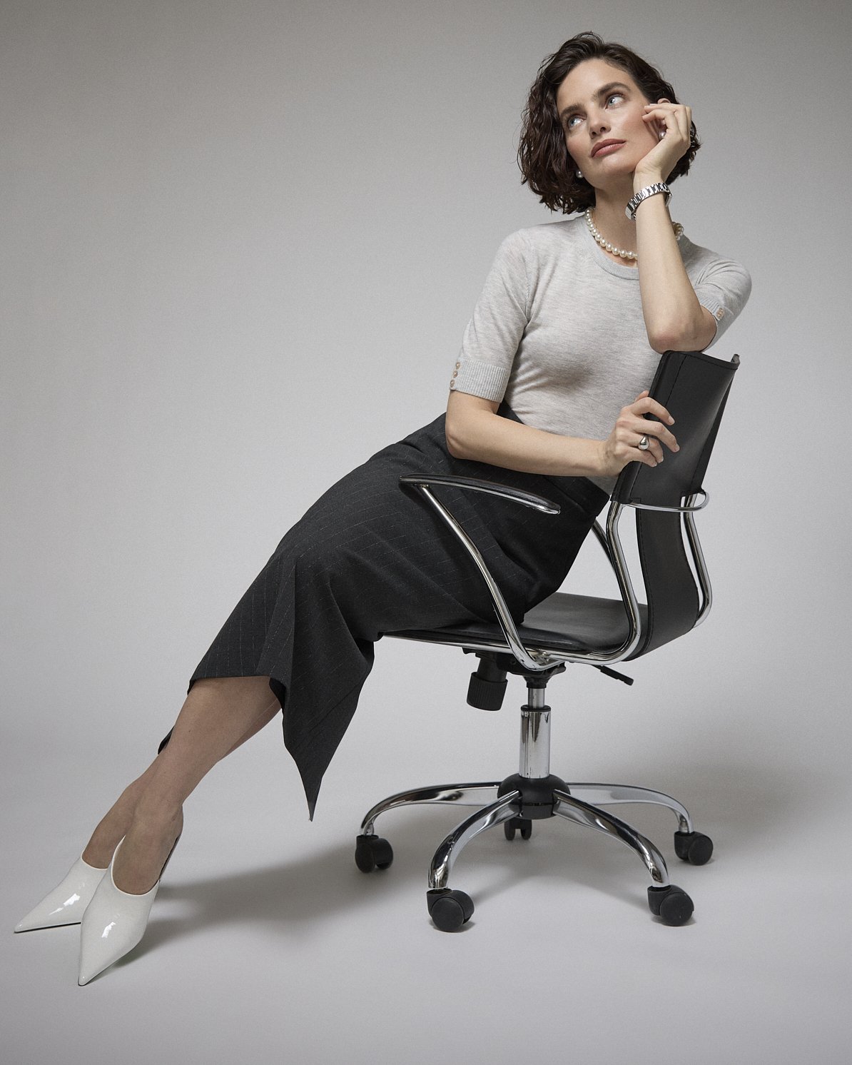 Woman in a grey knit top and black skirt leaning back on an office chair with a telephone.