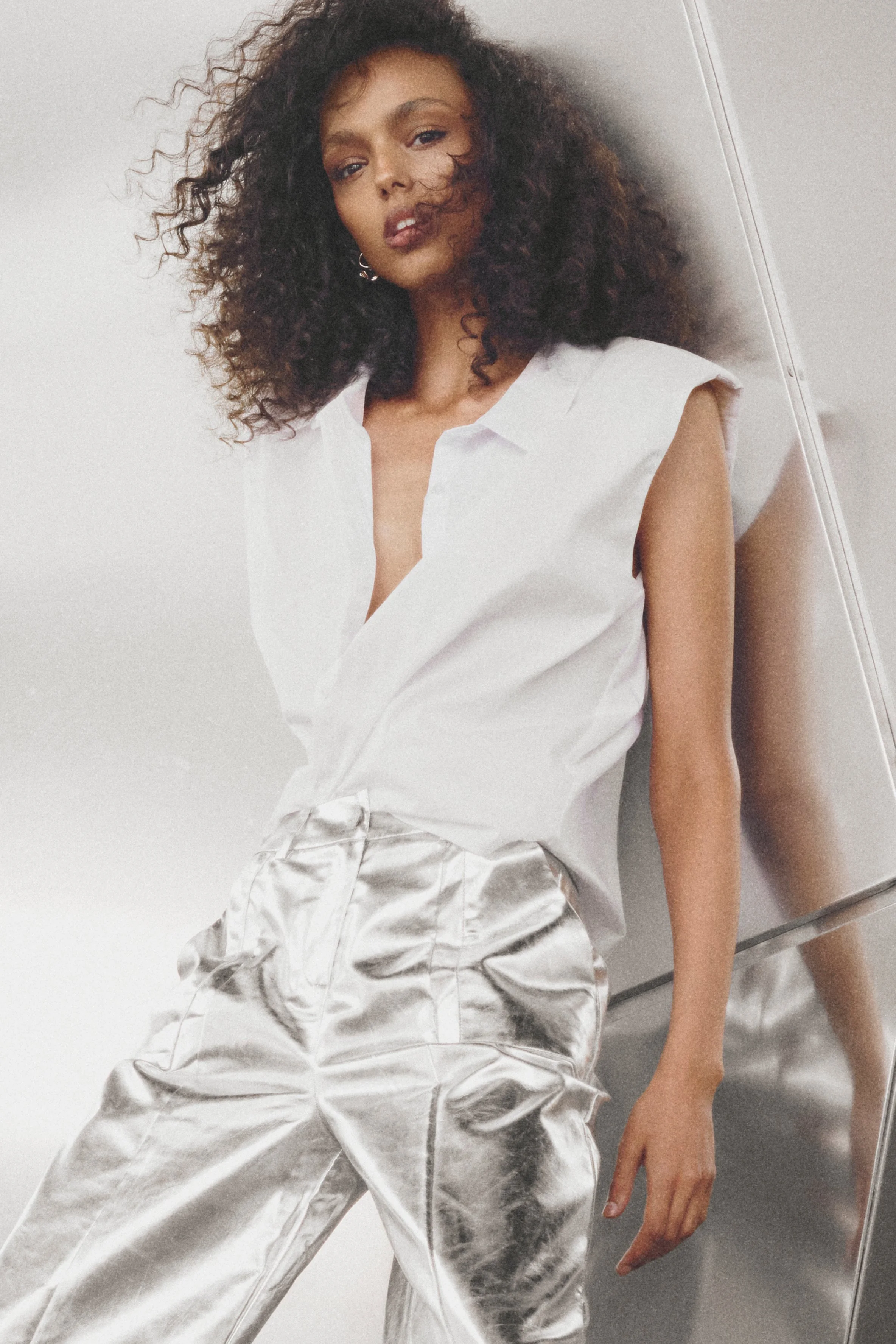 Woman in a white sleeveless blouse and metallic silver trousers against a curved metal wall.