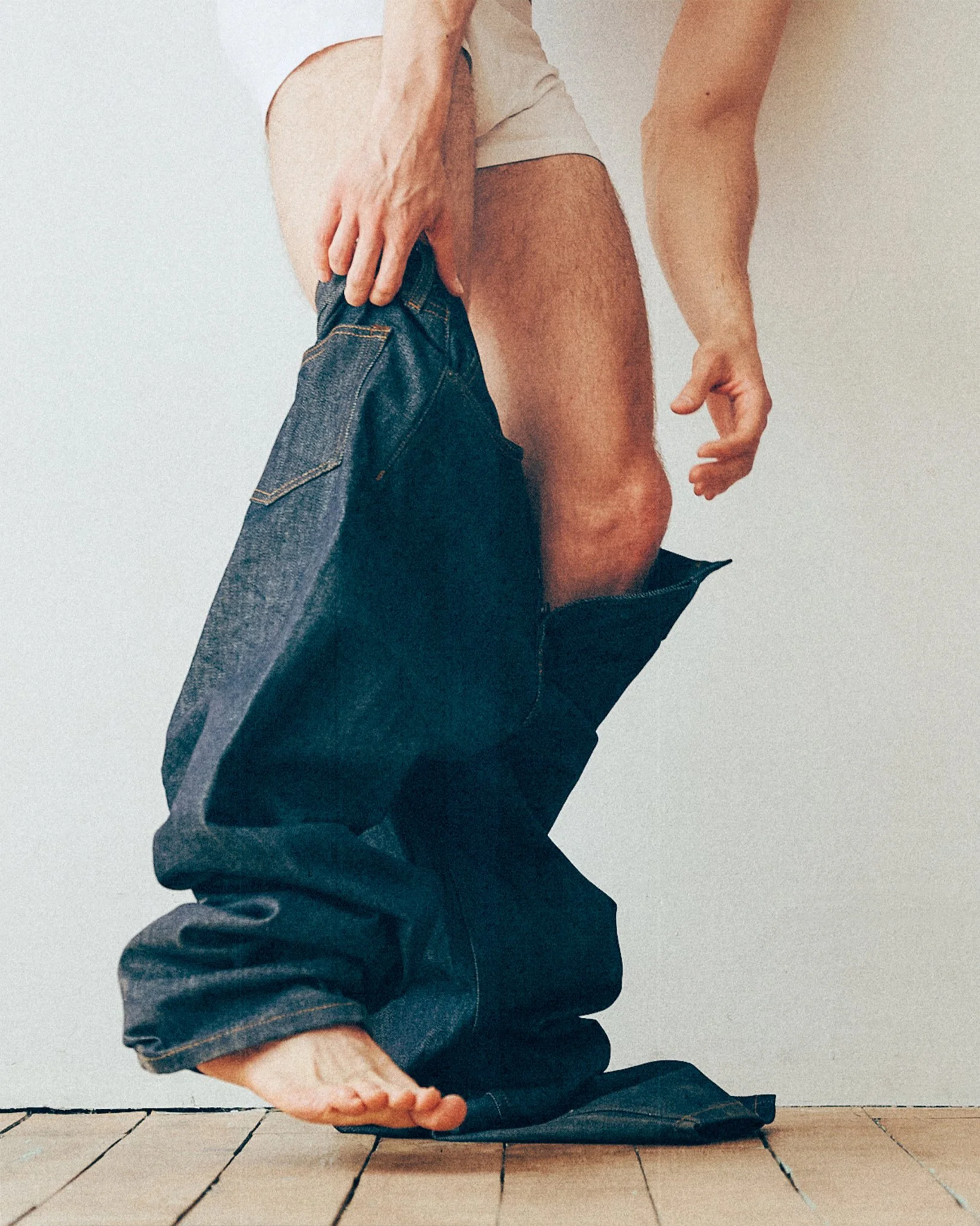 Detail of a man's legs and bare feet as he holds dark jeans below his white underwear.