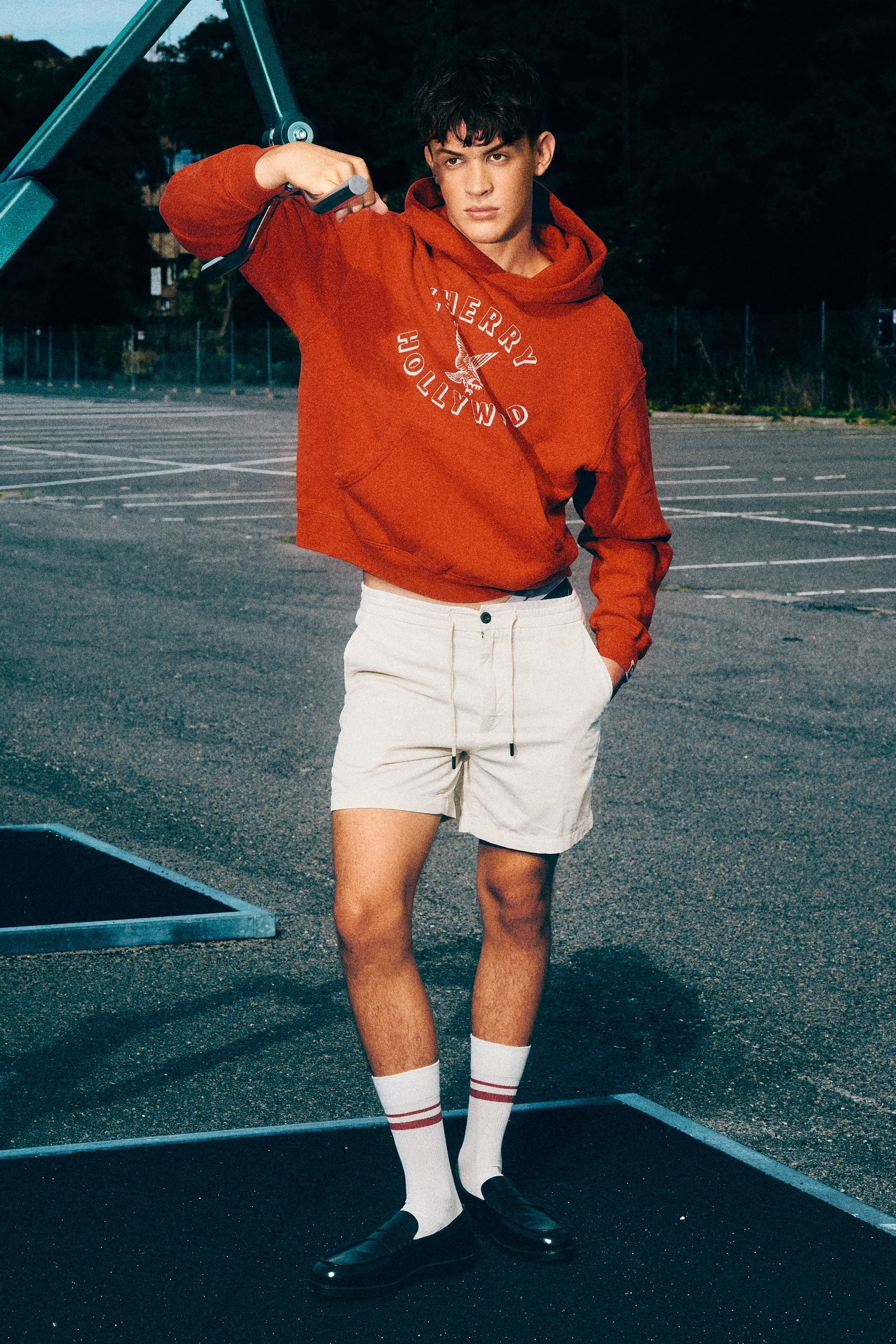 Man in a red sweatshirt and white shorts standing beside outdoor gym equipment.