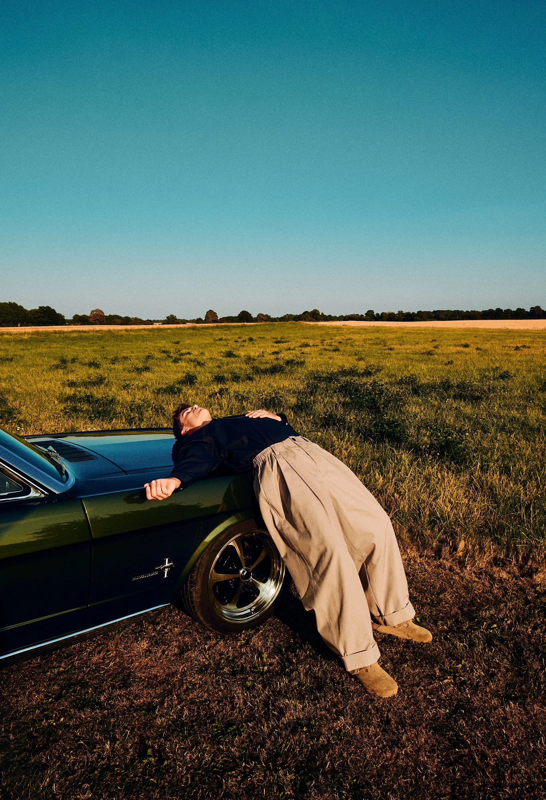 Man in a dark top and wide beige trousers leaning back against a convertible beneath a blue sky.