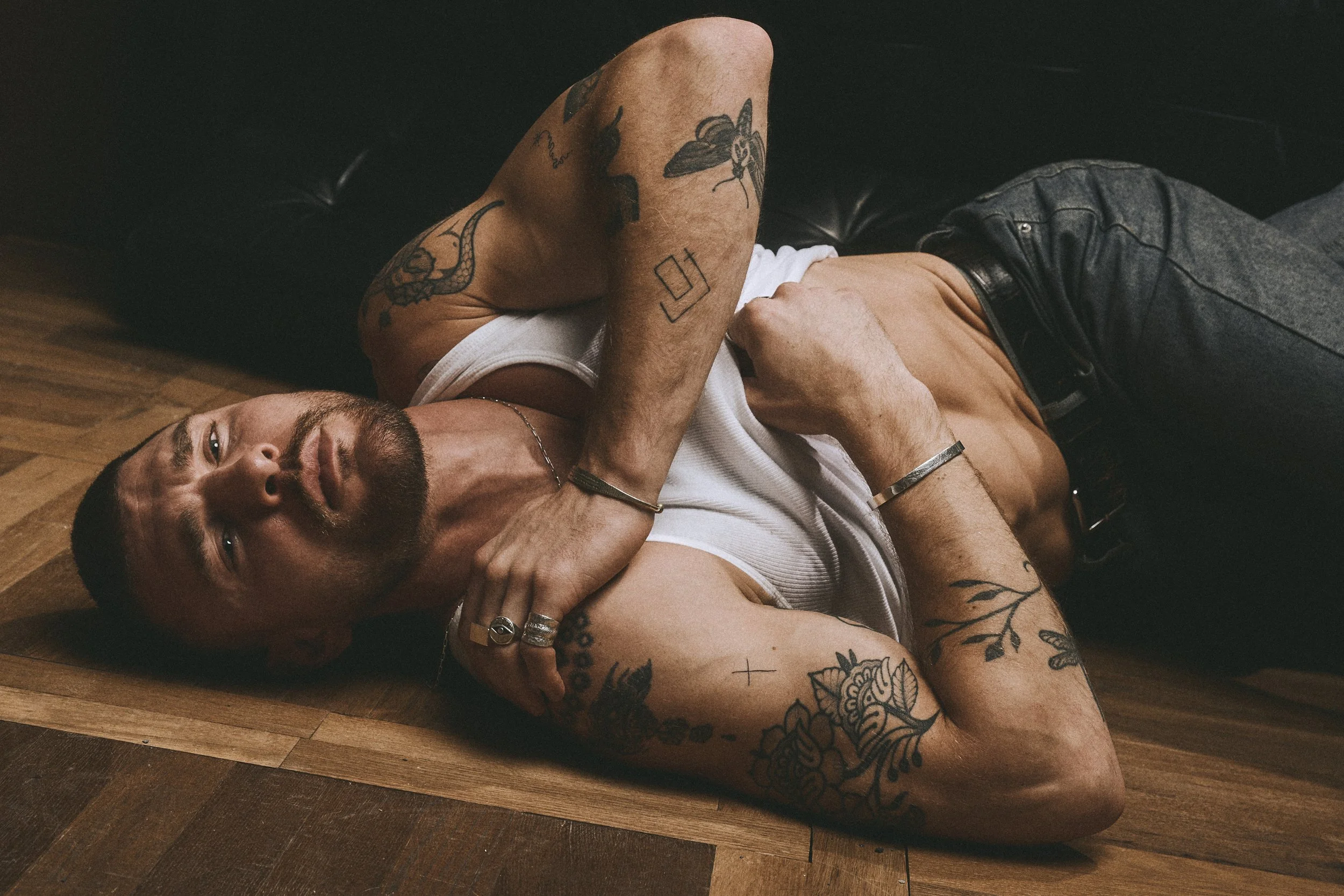 Close view of a tattooed man in a white tank top lying on a wooden floor with a hand across his chest.