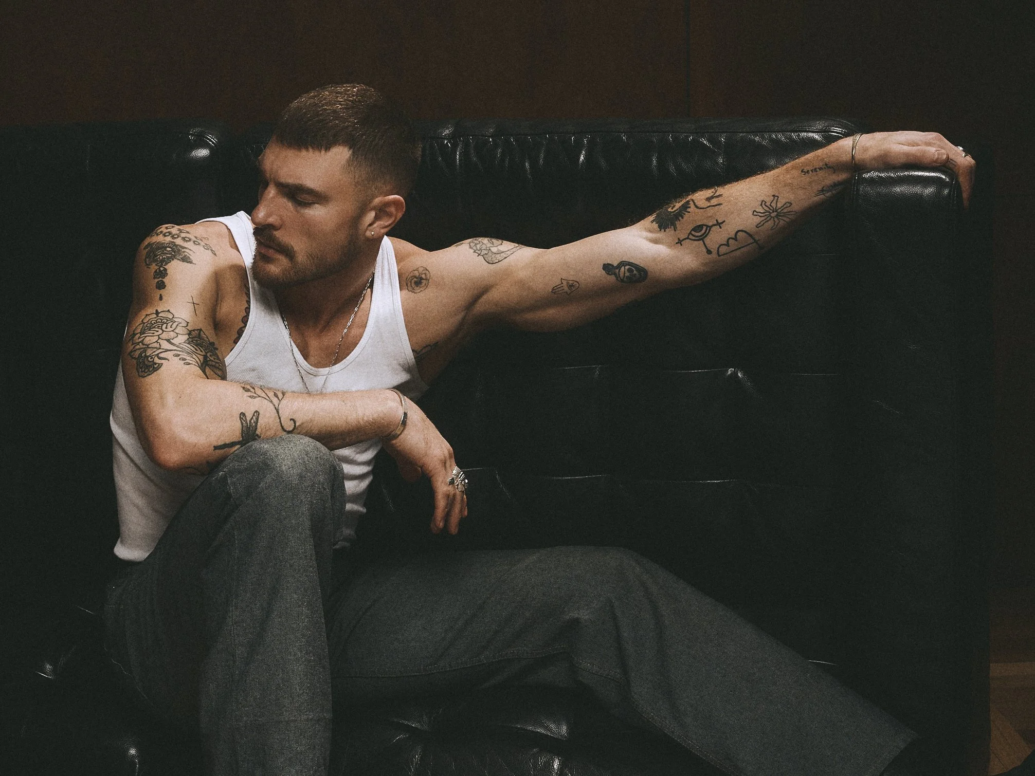 Tattooed man in a white tank top seated beside a leather sofa arm, looking to the side.