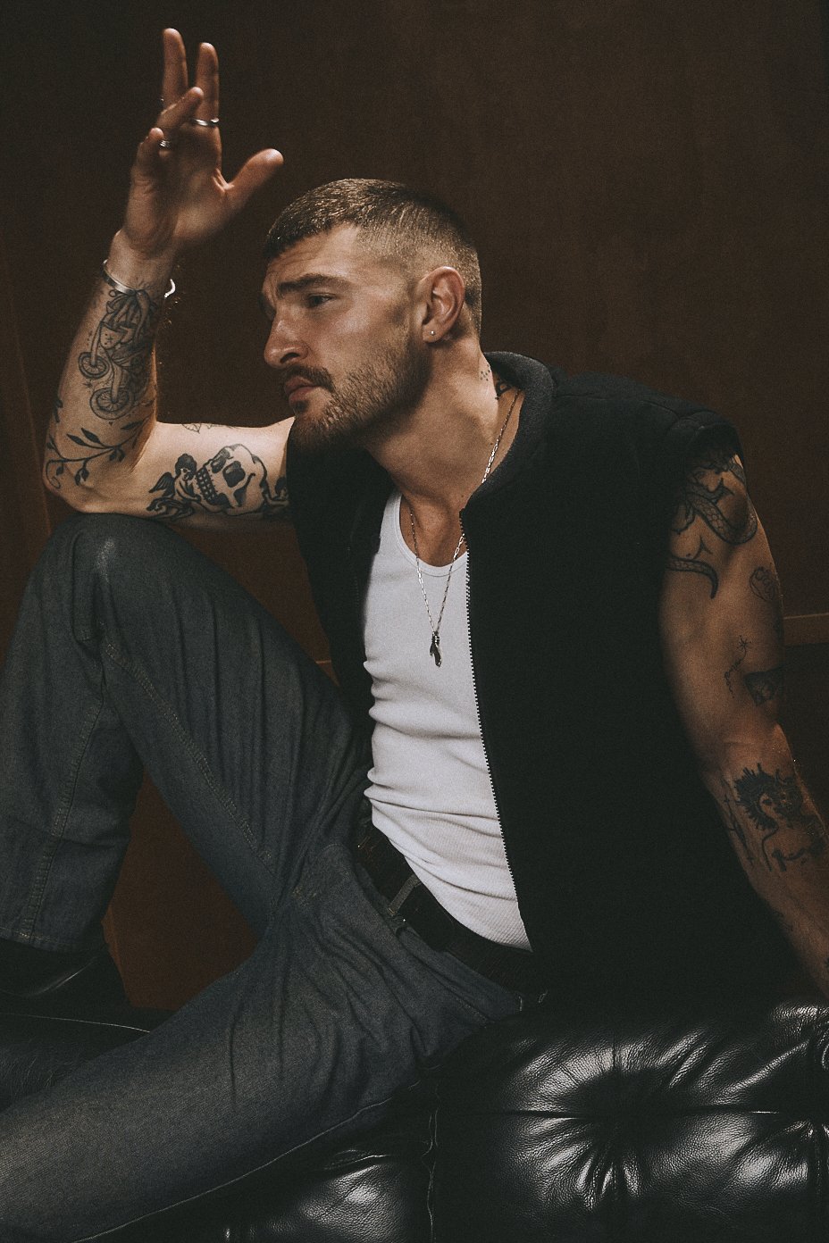 Tattooed man in a dark vest seated on a leather sofa with one hand raised above his head.