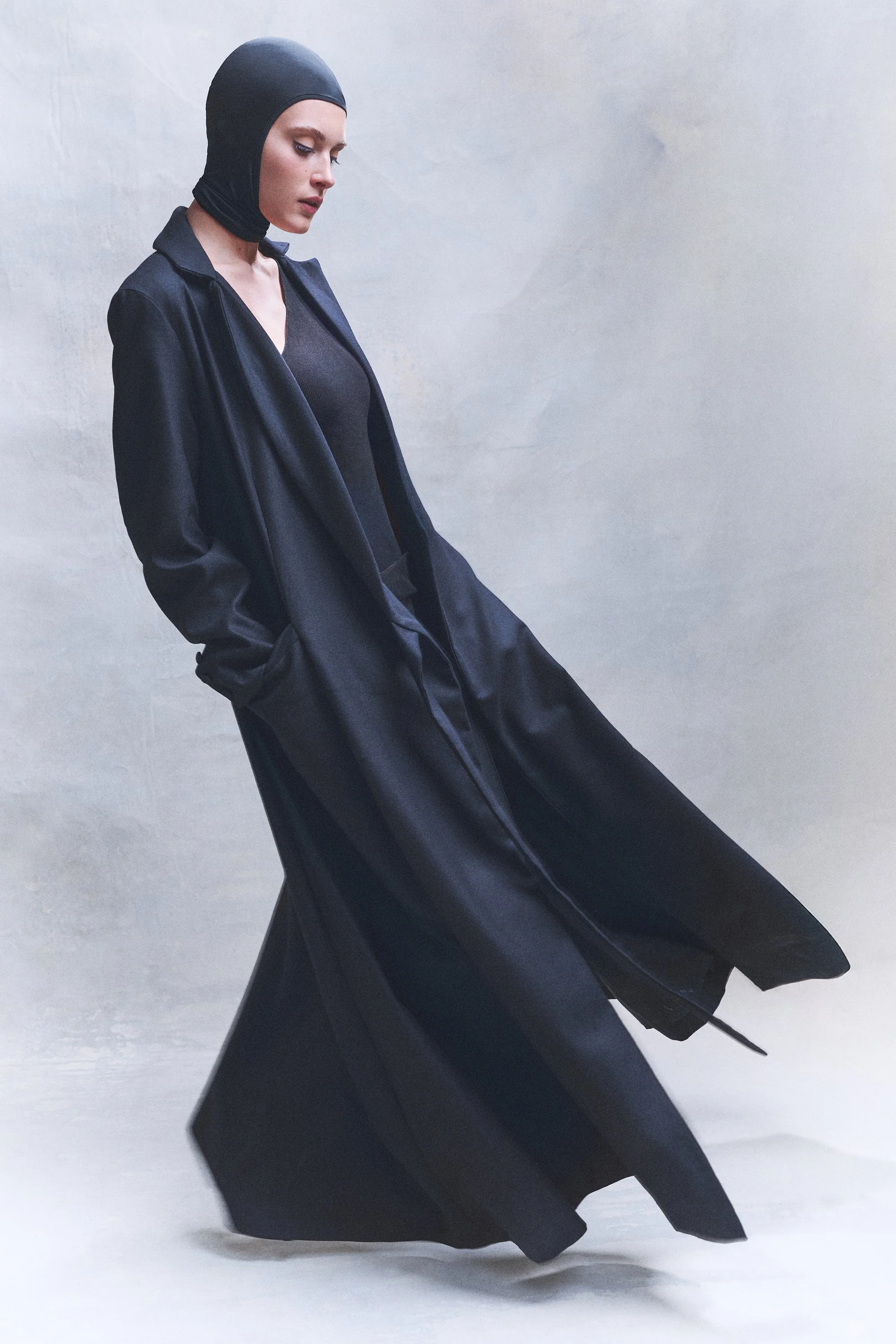 Woman in a flowing long black coat and close-fitting black head covering looking down in profile.