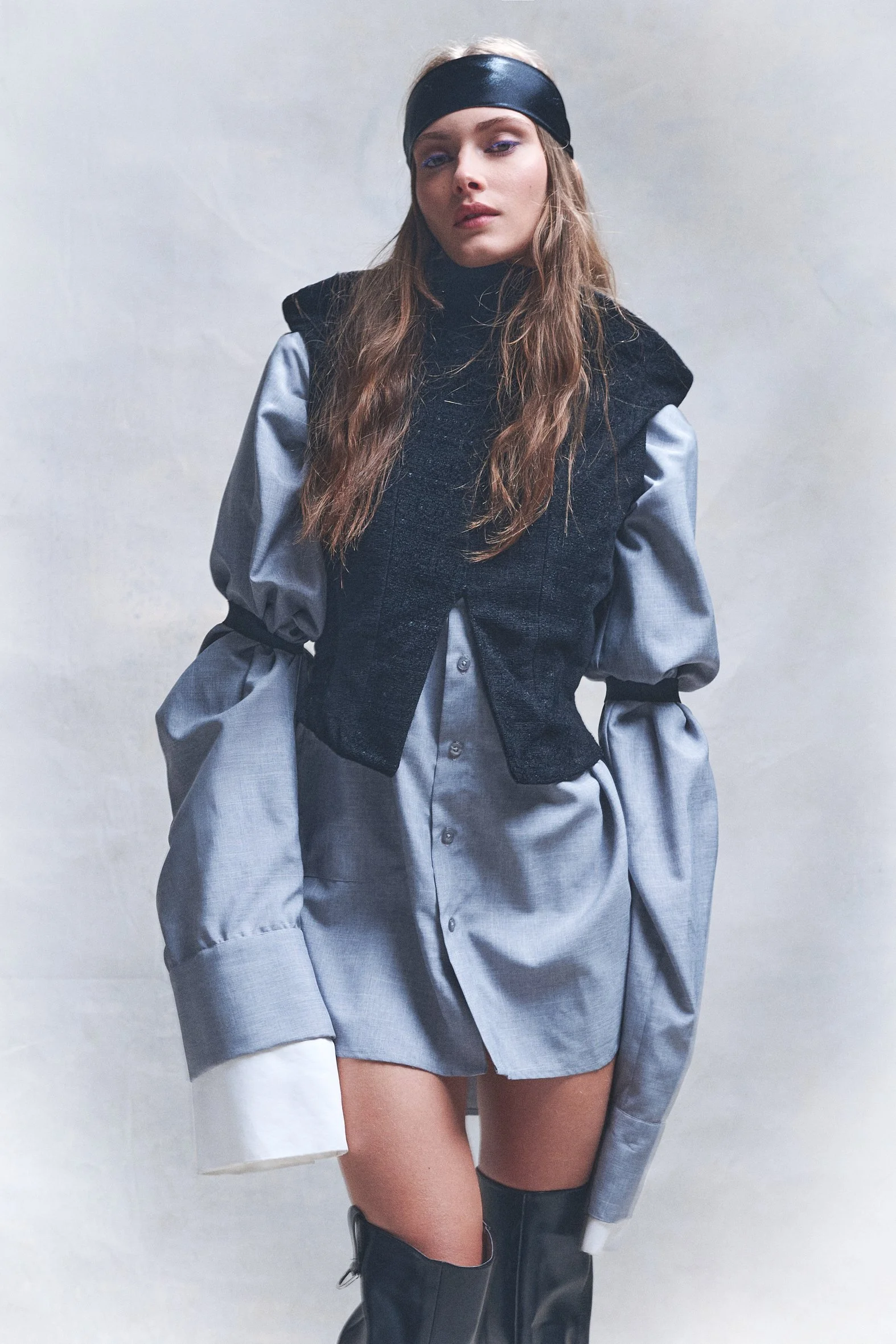 Woman in a silver oversized shirt, black vest and dark head covering against a pale studio backdrop.