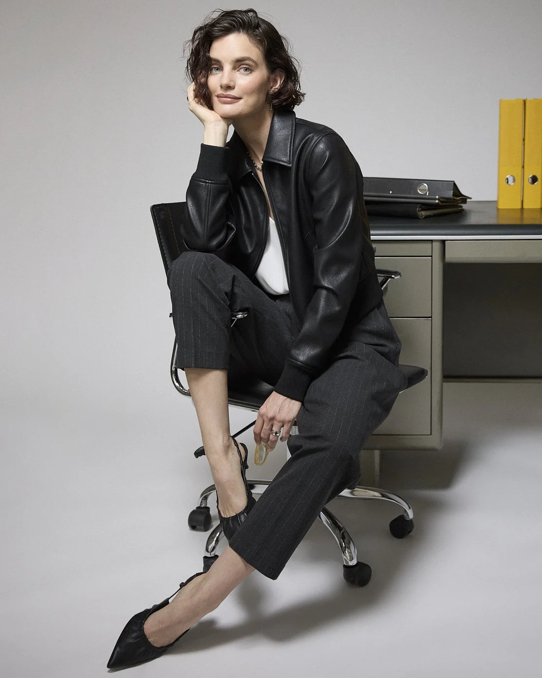 Woman in a black leather jacket and trousers seated on an office chair beside filing cabinets.