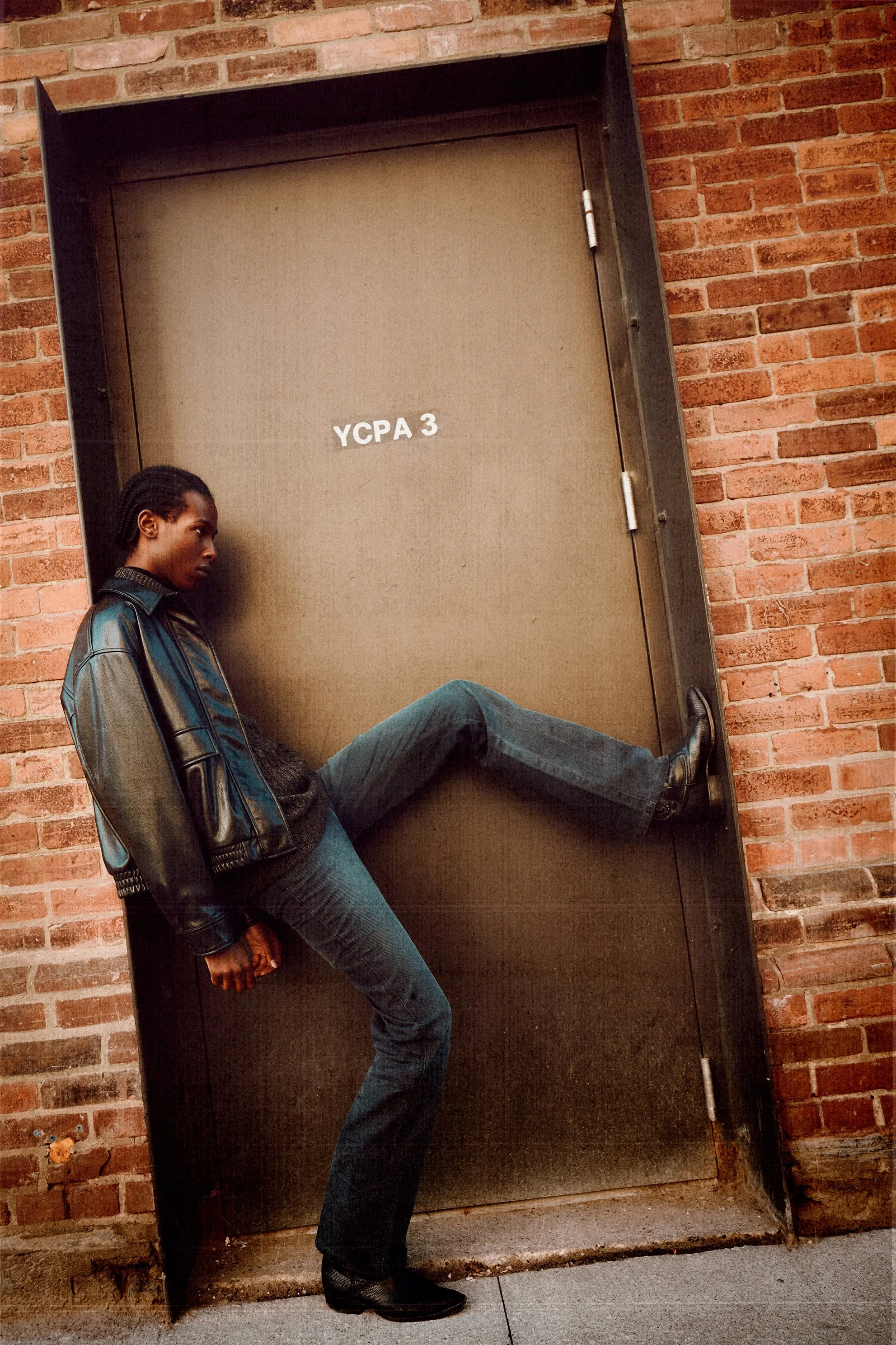 Man in a leather jacket and jeans leaning against an industrial door with one leg raised.