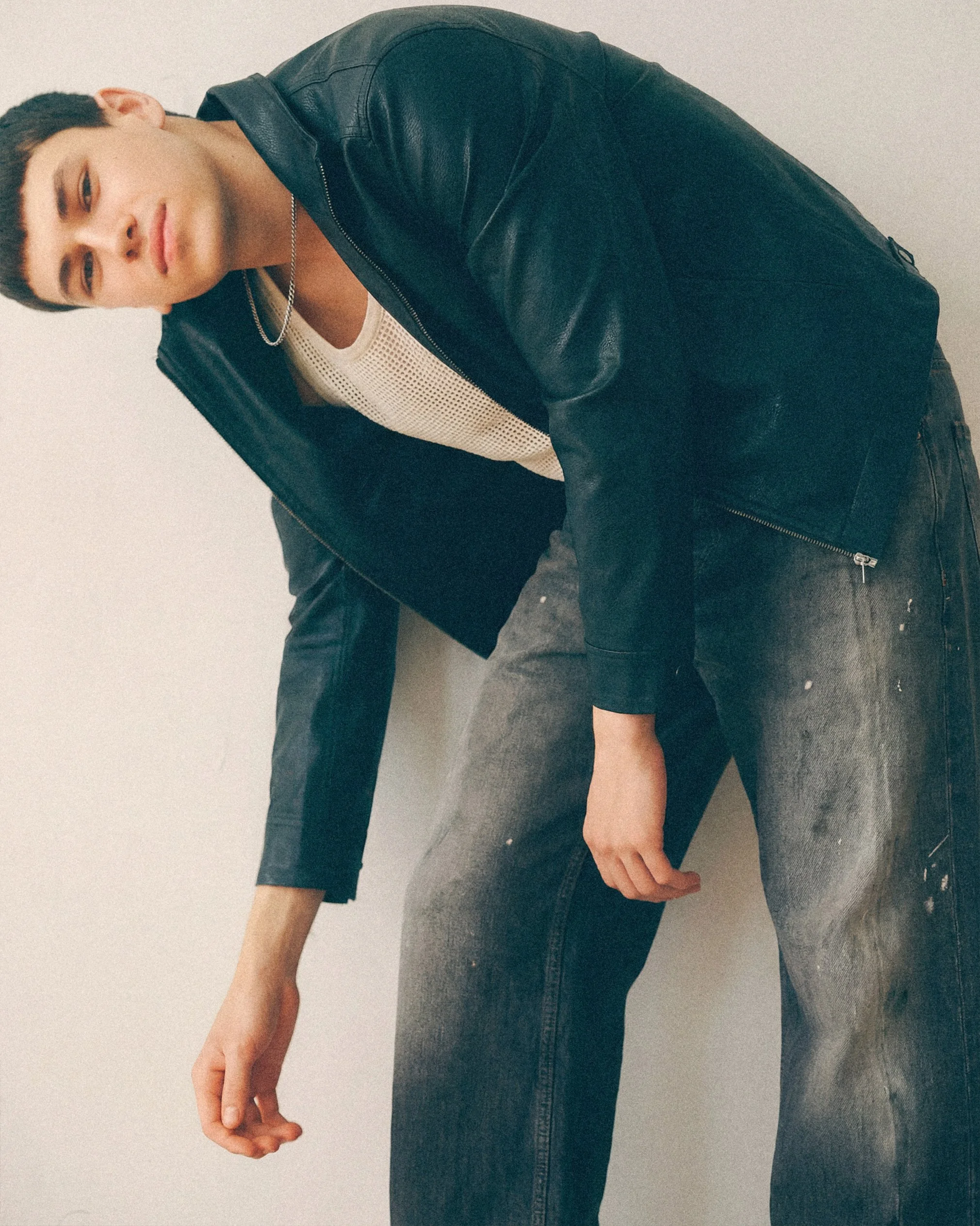 Young man bending forward in a black jacket and jeans with his arms hanging loosely.