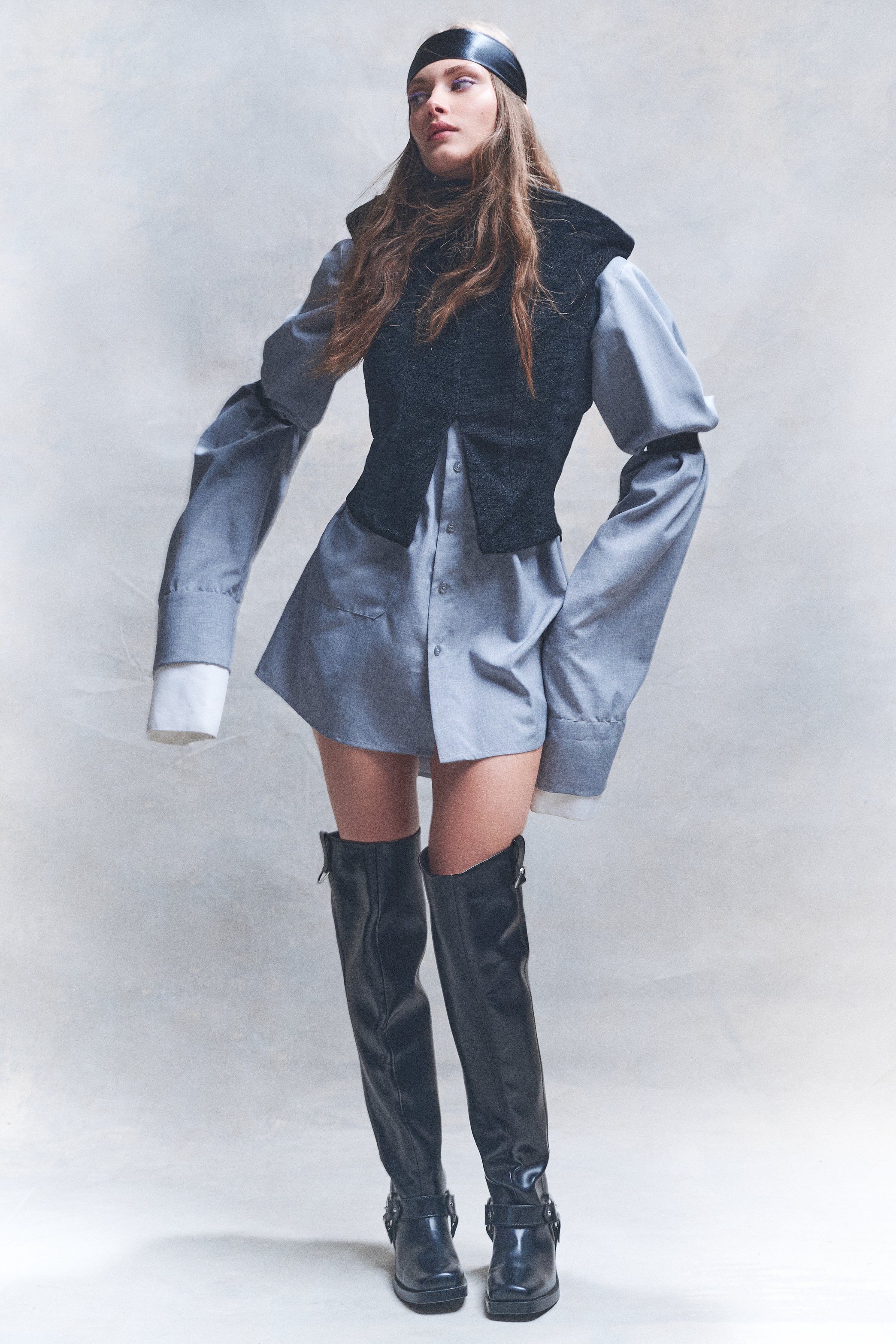 Full-length portrait of a woman in an oversized silver shirt, black vest and knee-high black boots.