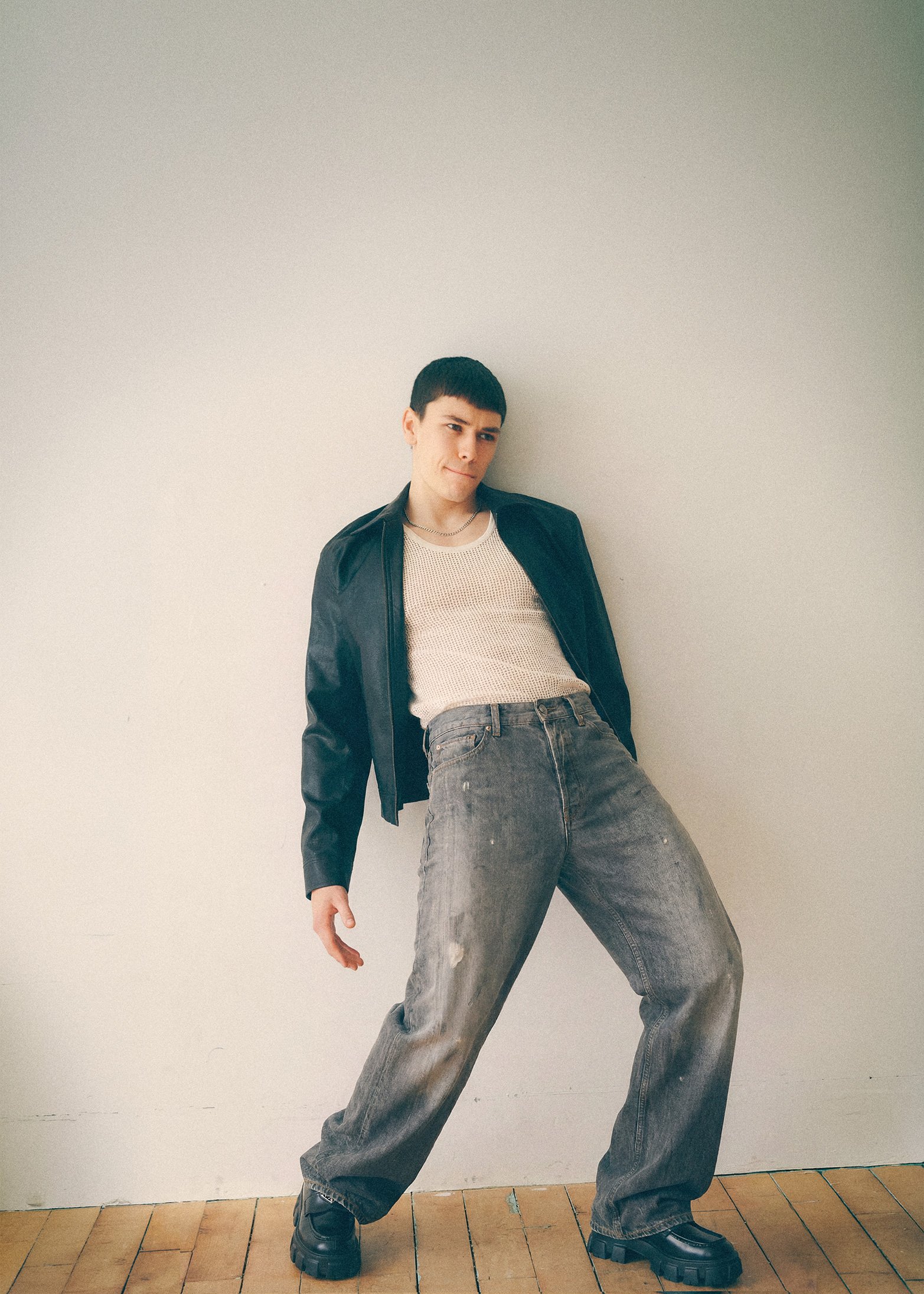 Young man standing in a black jacket, white tank top and loose jeans against a pale wall.