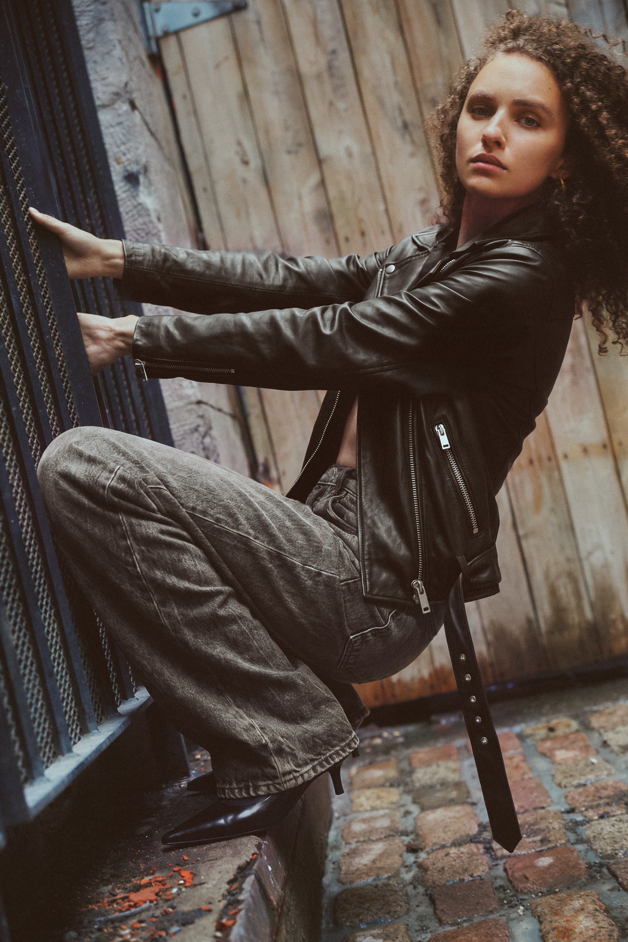 Fashion model in a black leather jacket and faded grey jeans posing against an iron gate in a cobblestone alley.