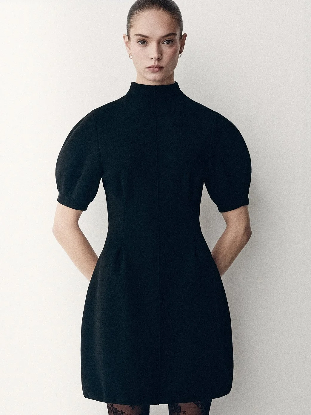 Woman facing the camera in a short black dress with puff sleeves, hands behind her back.
